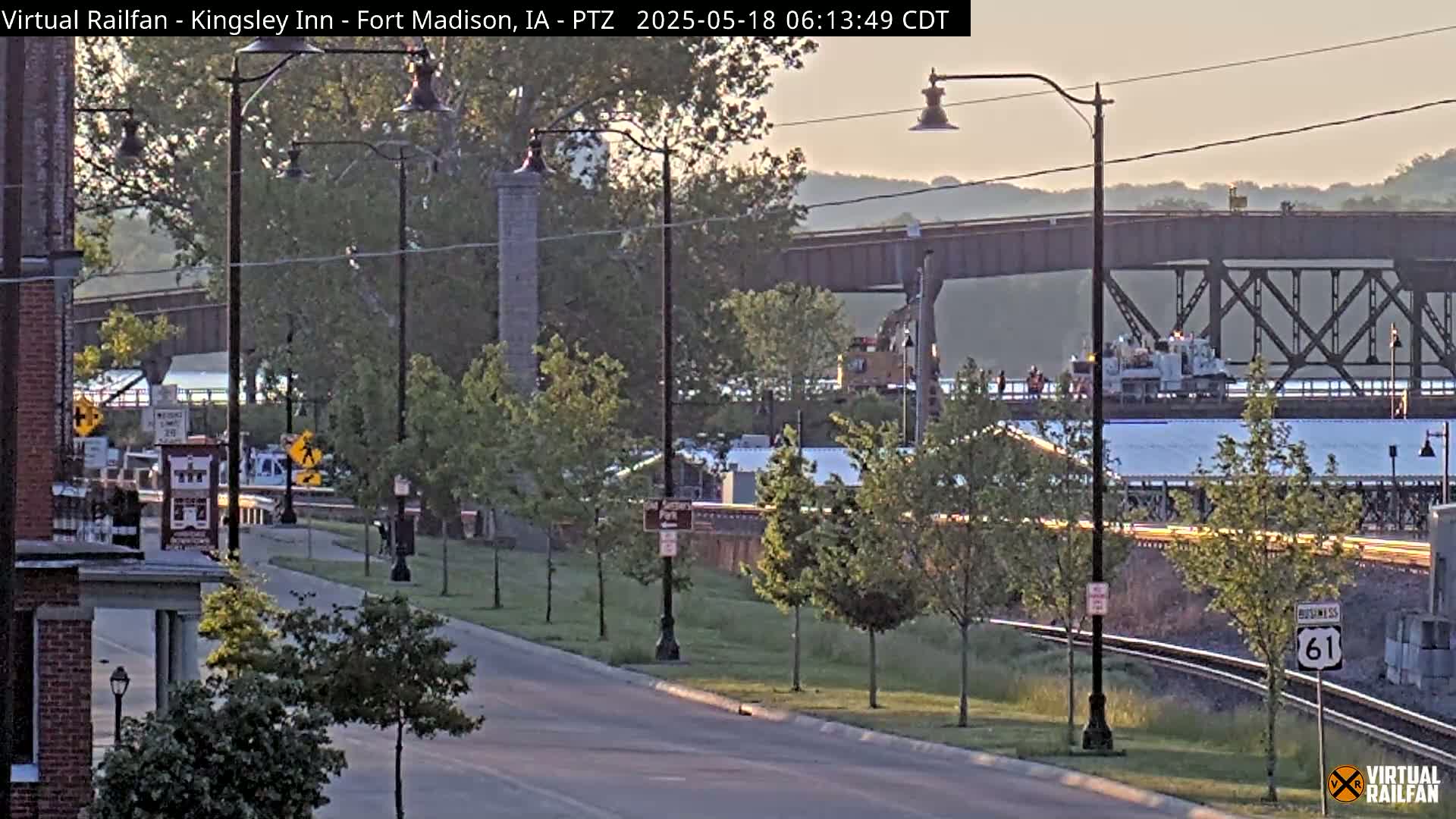 A view of a street with lampposts and trees, alongside a railway line and a metal bridge over a body of water, under a partly cloudy, soft light sky at dawn.