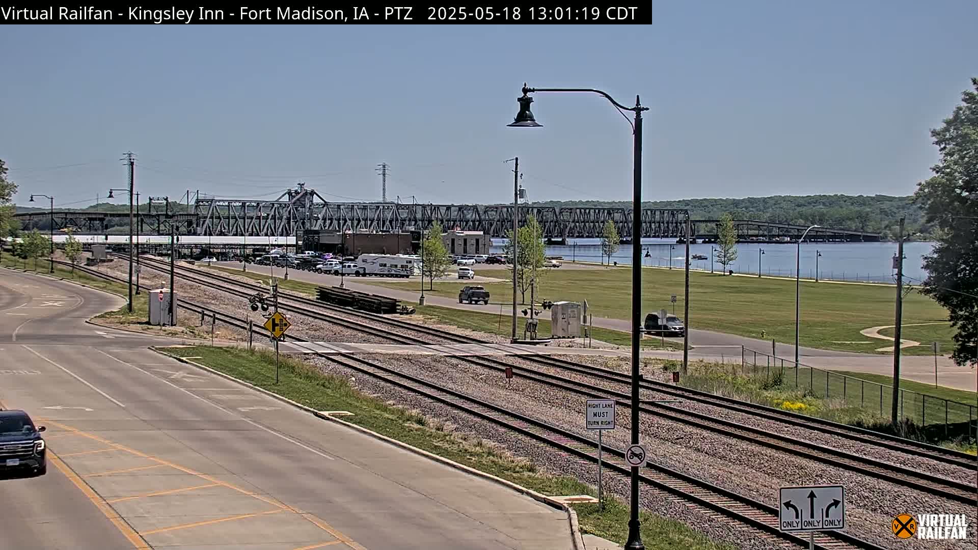 A sunny day shows a view of railroad tracks intersecting with a road, leading to a bridge spanning a river with a park and a few vehicles visible.
