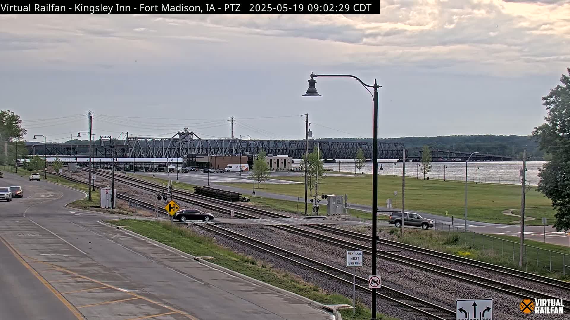 A mostly cloudy day reveals a view of railroad tracks intersecting a road, with a river and large bridge in the background.
