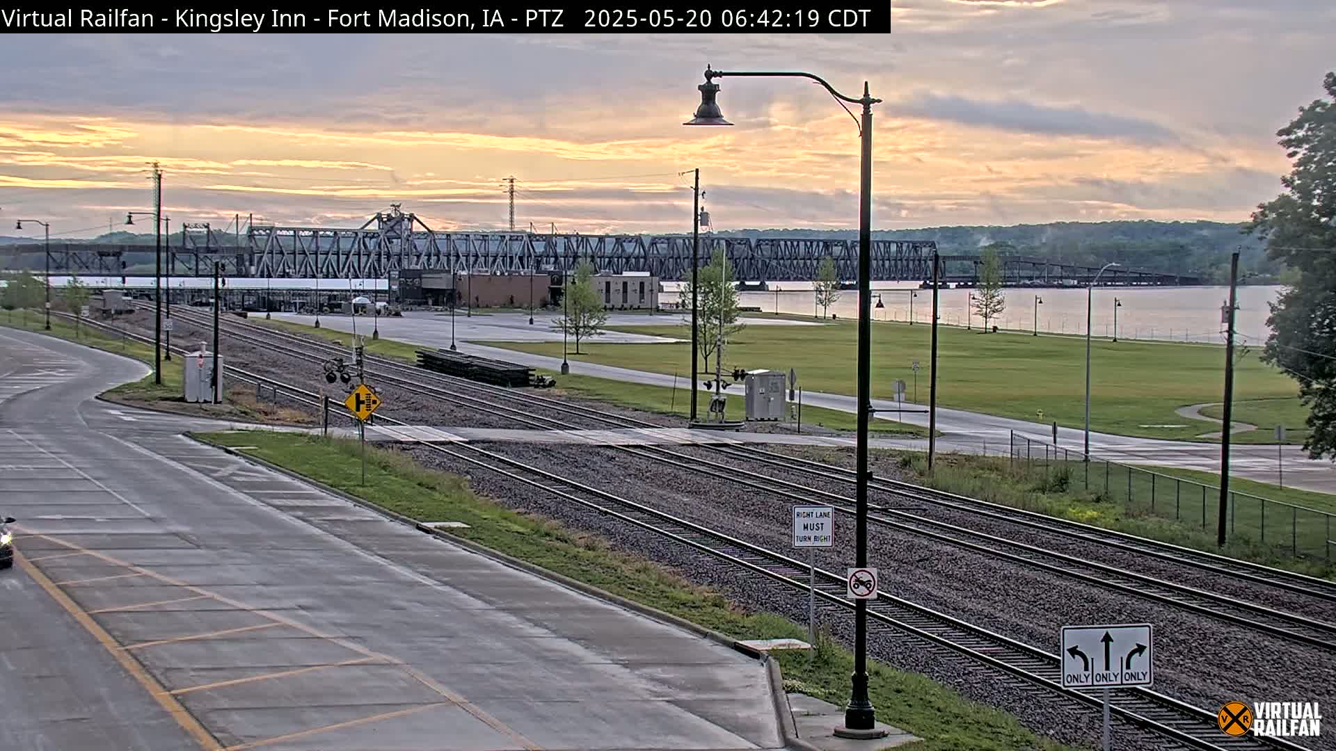 A sunrise scene shows railroad tracks intersecting a road in front of a river and a large metal bridge under a partly cloudy sky.