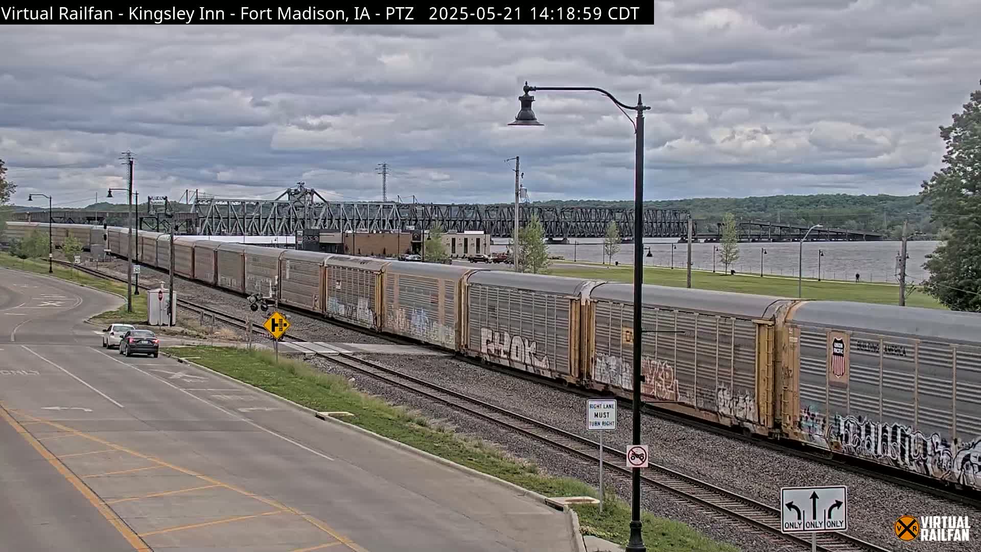 A long freight train passes by a road and under a bridge on an overcast day near a river.