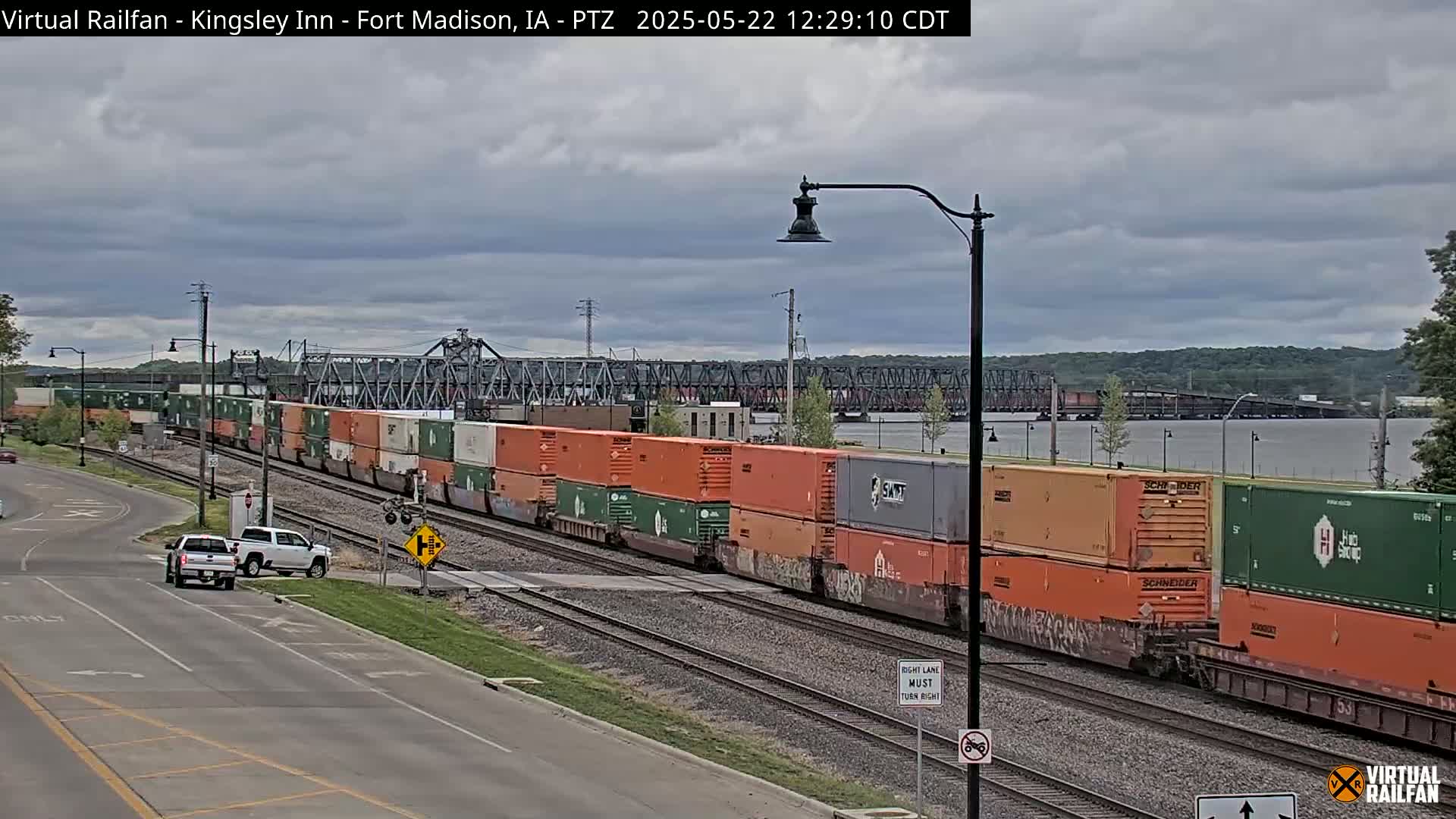 A long freight train carrying numerous orange and green shipping containers passes a road under an overcast sky, with a river and bridge visible in the background.