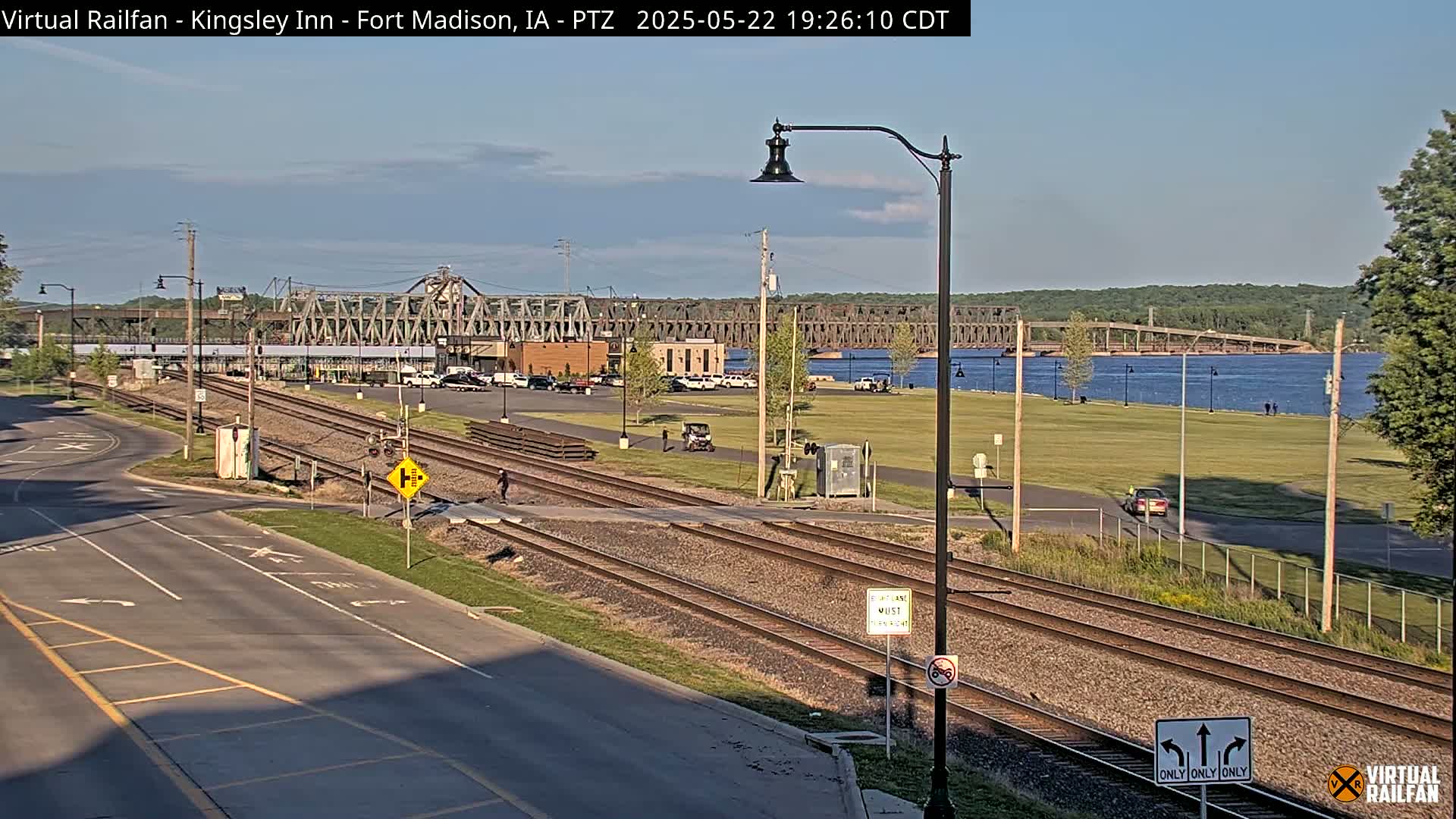A view of railroad tracks intersecting a road, with a large bridge and river in the background under a clear sky.