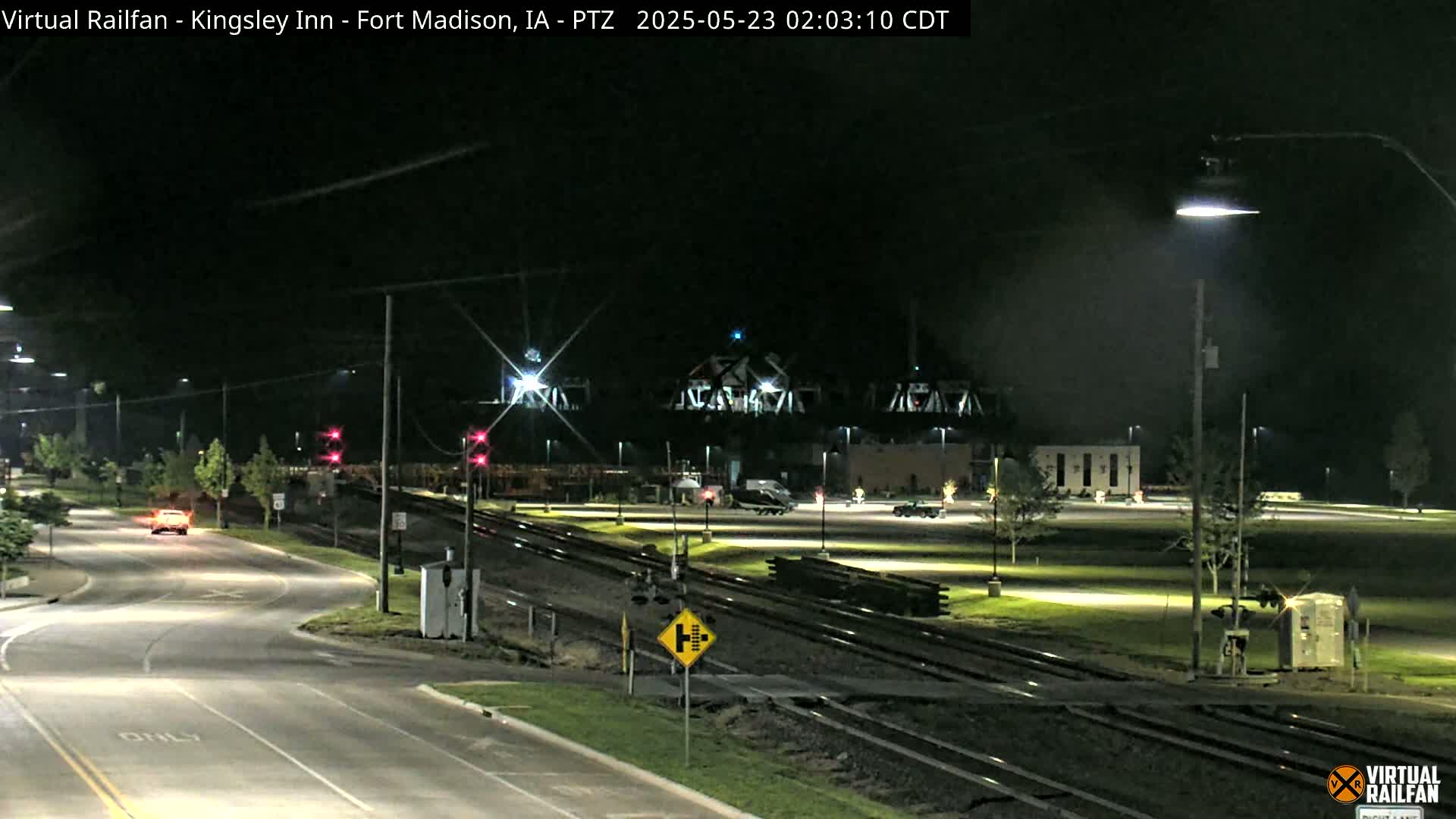 A nighttime view of a road intersection near railroad tracks and a bridge, with a single car visible on the road.