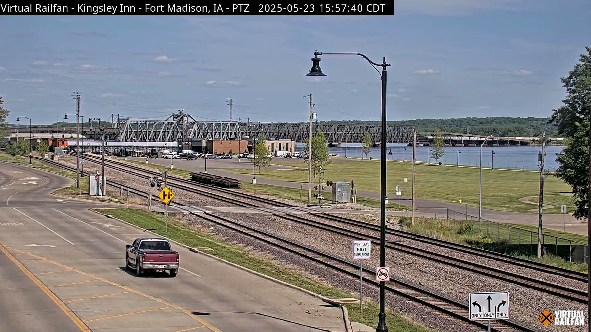 A red pickup truck drives on a road next to train tracks and a river, with a metal bridge in the background under a partly cloudy sky.
