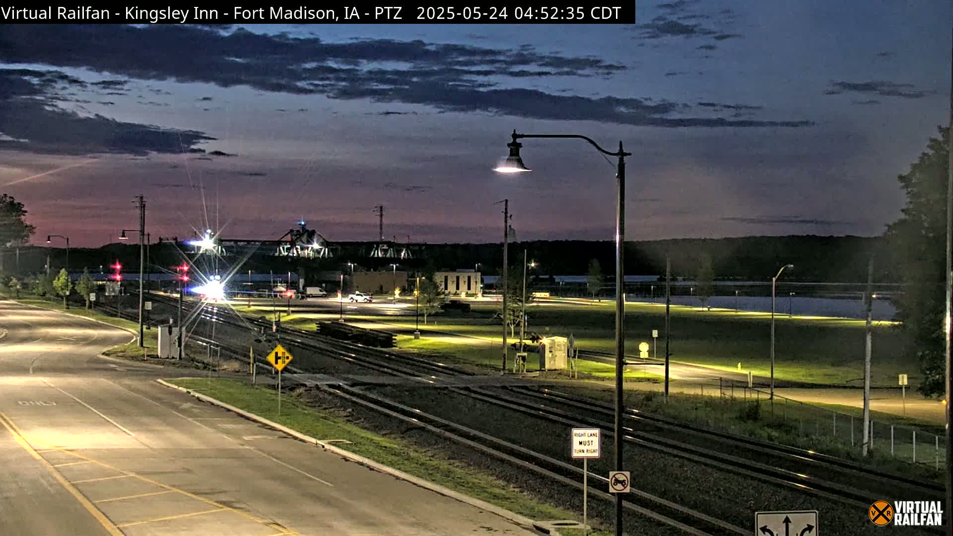 A twilight view of a train crossing near a body of water, with a roadway and train tracks visible, under a mostly clear sky.