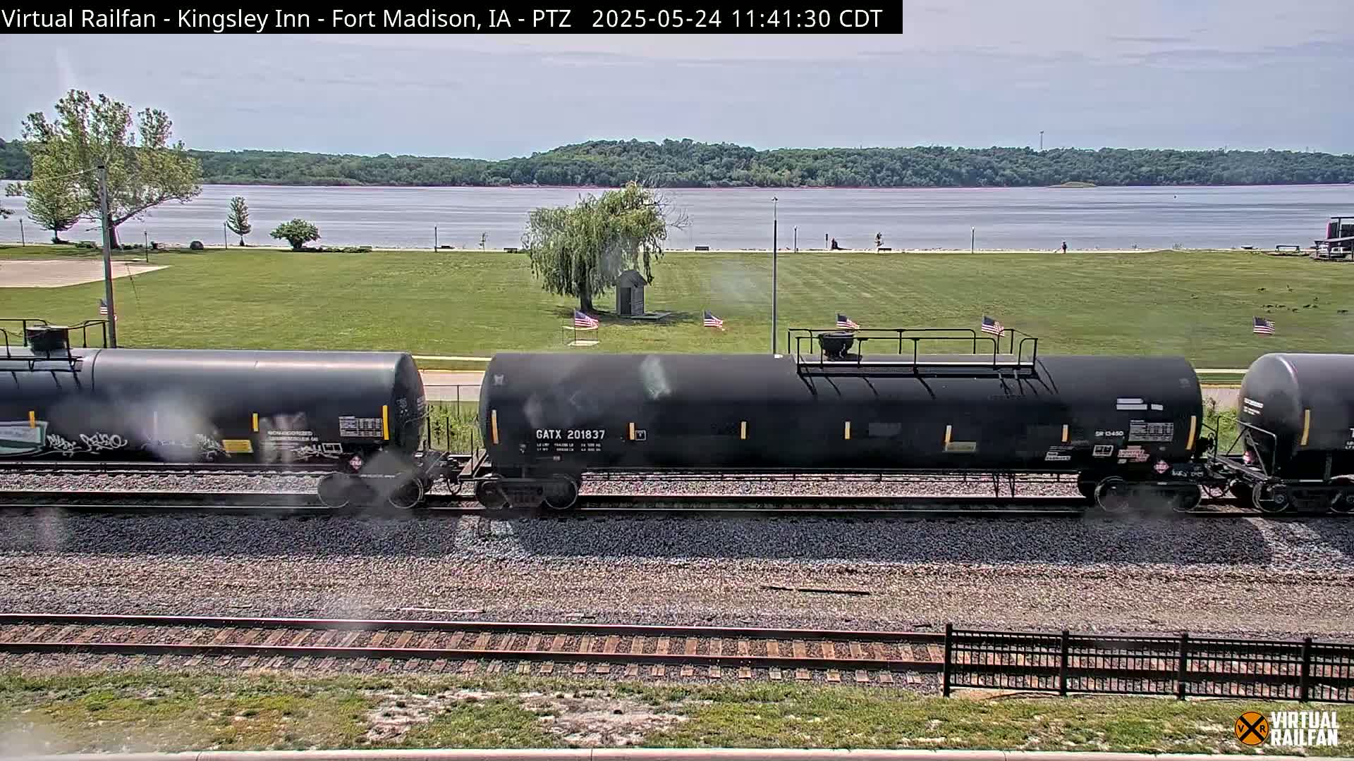 A freight train of black tanker cars sits on railroad tracks in front of a grassy area with a body of water and trees in the background under a mostly clear sky.