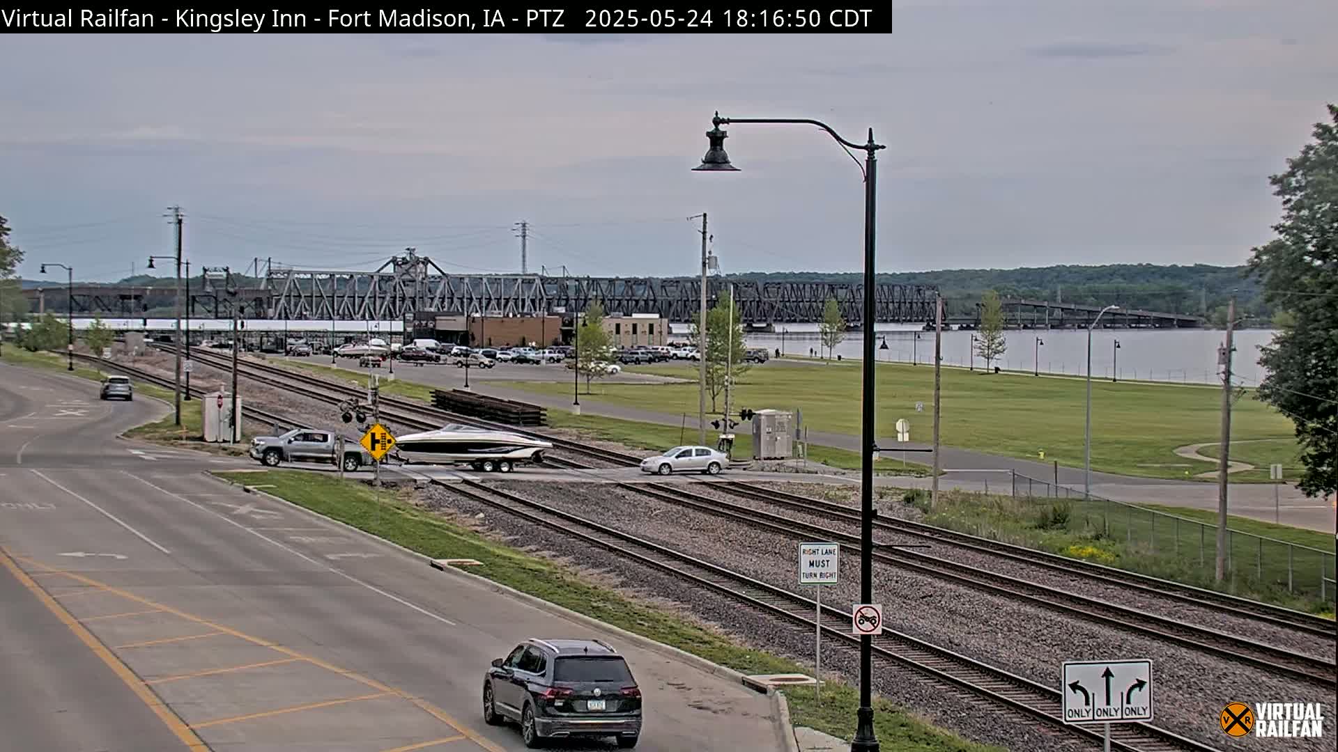 A partly cloudy day reveals a railroad crossing with several vehicles, a boat on a trailer, and a large metal bridge in the background over a body of water.