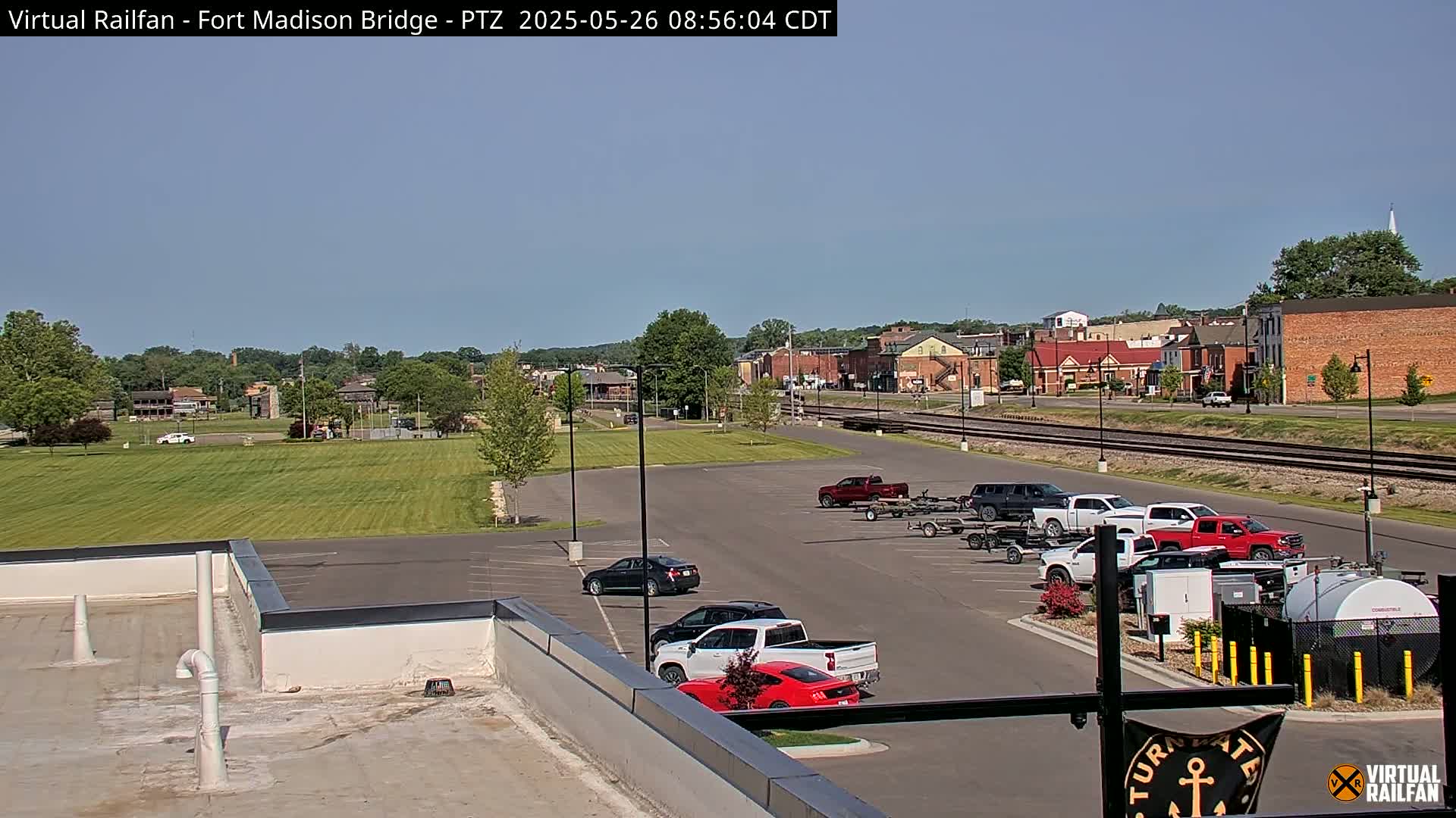 A mostly sunny day reveals a parking lot with several vehicles, a grassy area, railroad tracks, and a small town in the background.