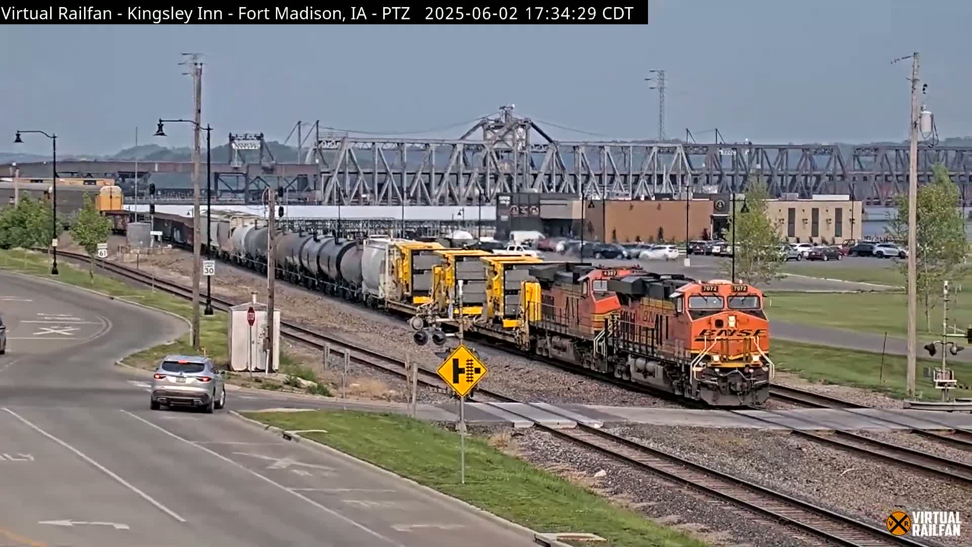 A long freight train, led by an orange locomotive, crosses a railroad crossing on a partly cloudy day, with a bridge and some buildings visible in the background.