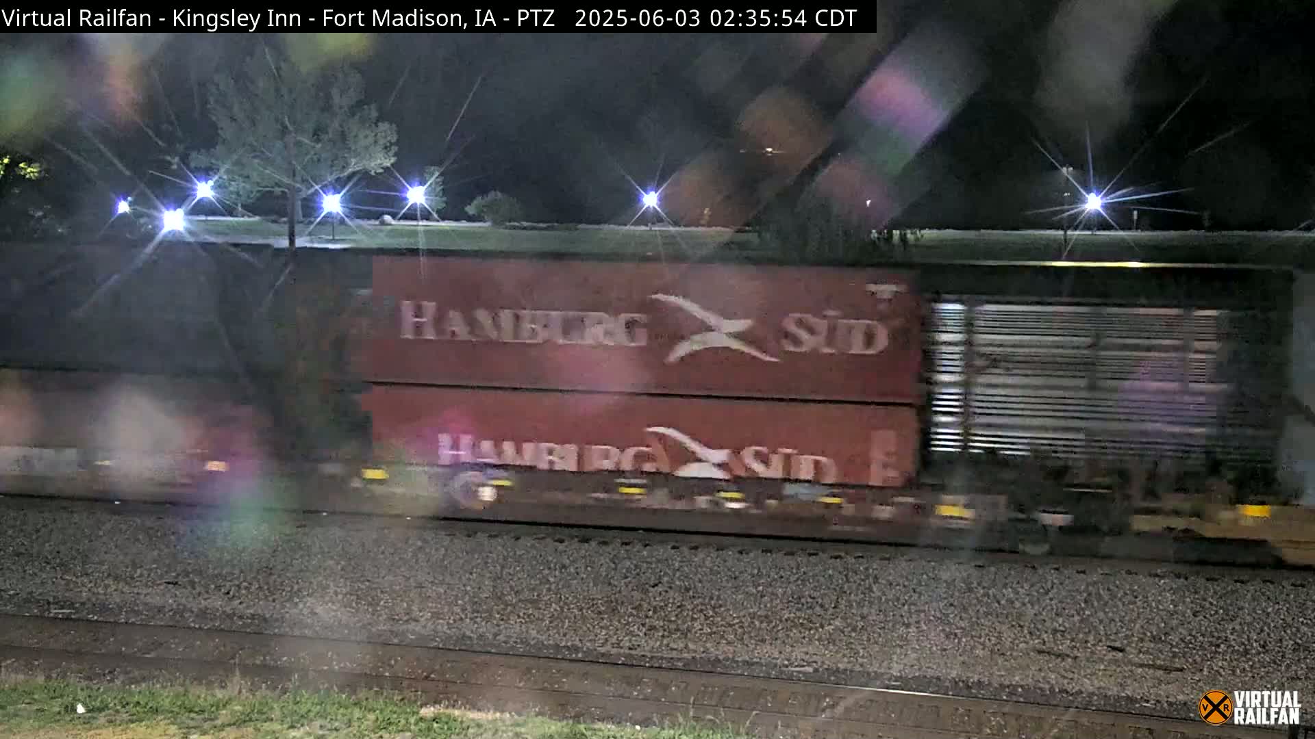 A freight train with red cars marked "Hamburg Süd" passes by at night under slightly wet conditions.