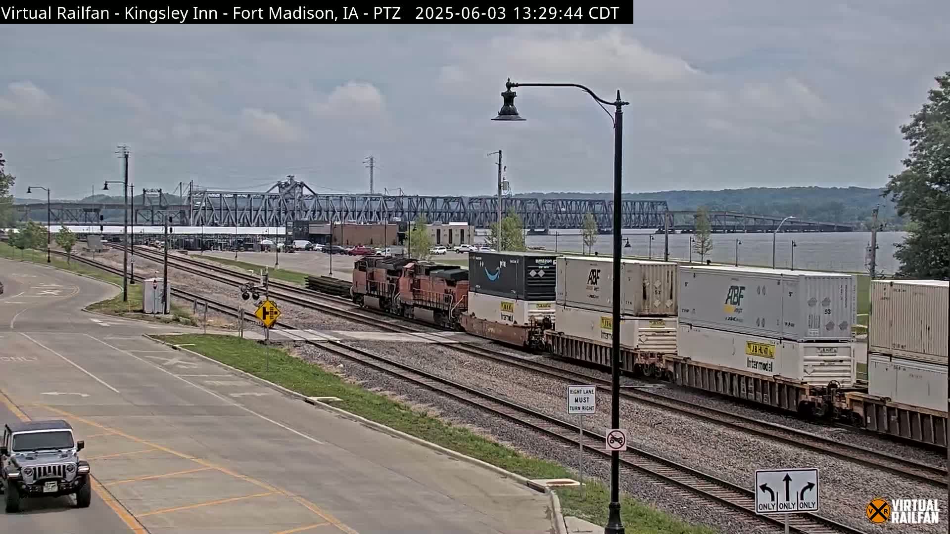 A freight train carrying shipping containers passes by a road and a river, under partly cloudy skies, with a bridge in the distance.