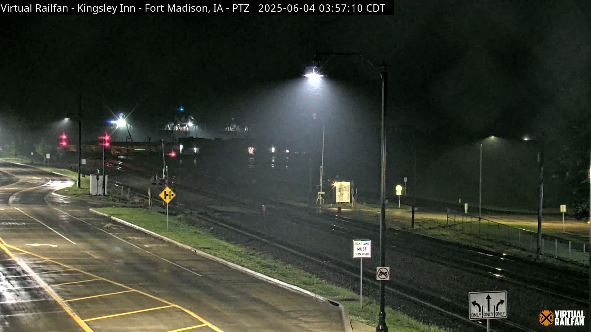 A wet, nighttime view of a road intersecting with railroad tracks, illuminated by streetlights under a dark, overcast sky.