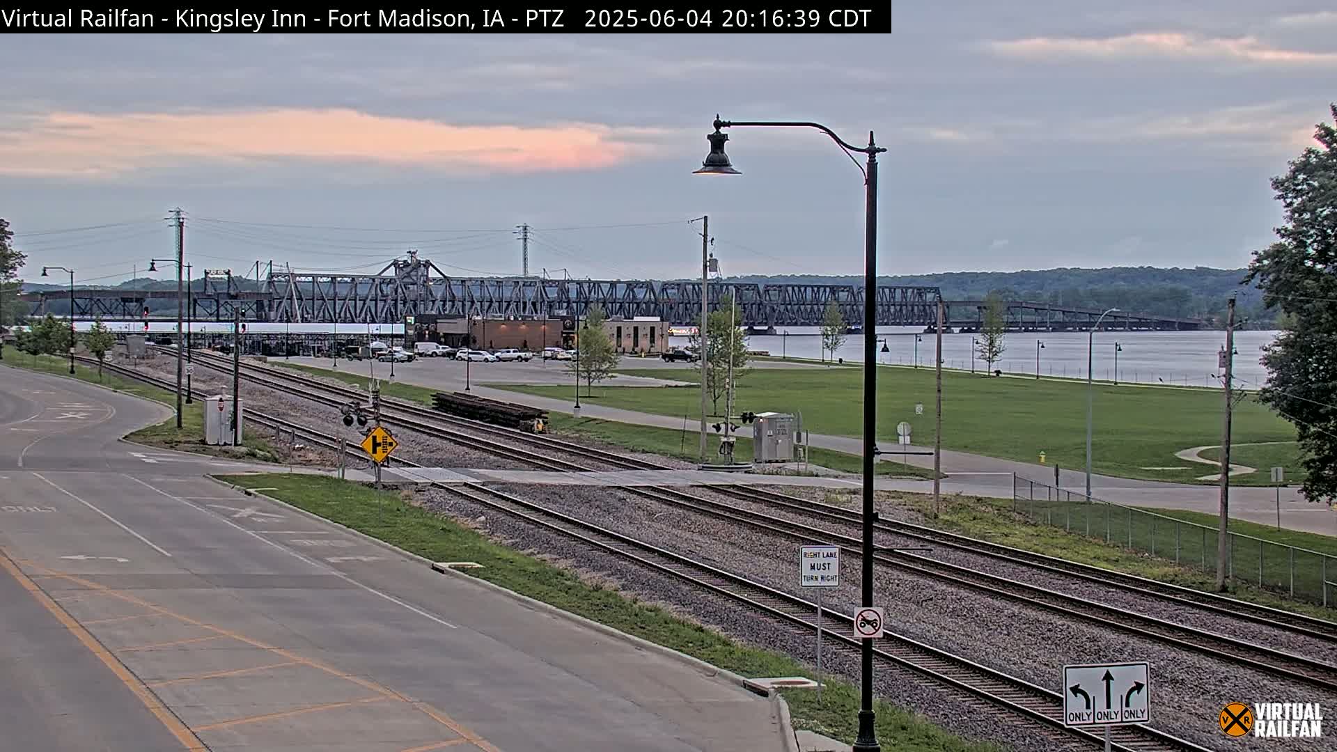Under an overcast sky, railroad tracks cross a road in front of a river and a large metal bridge.