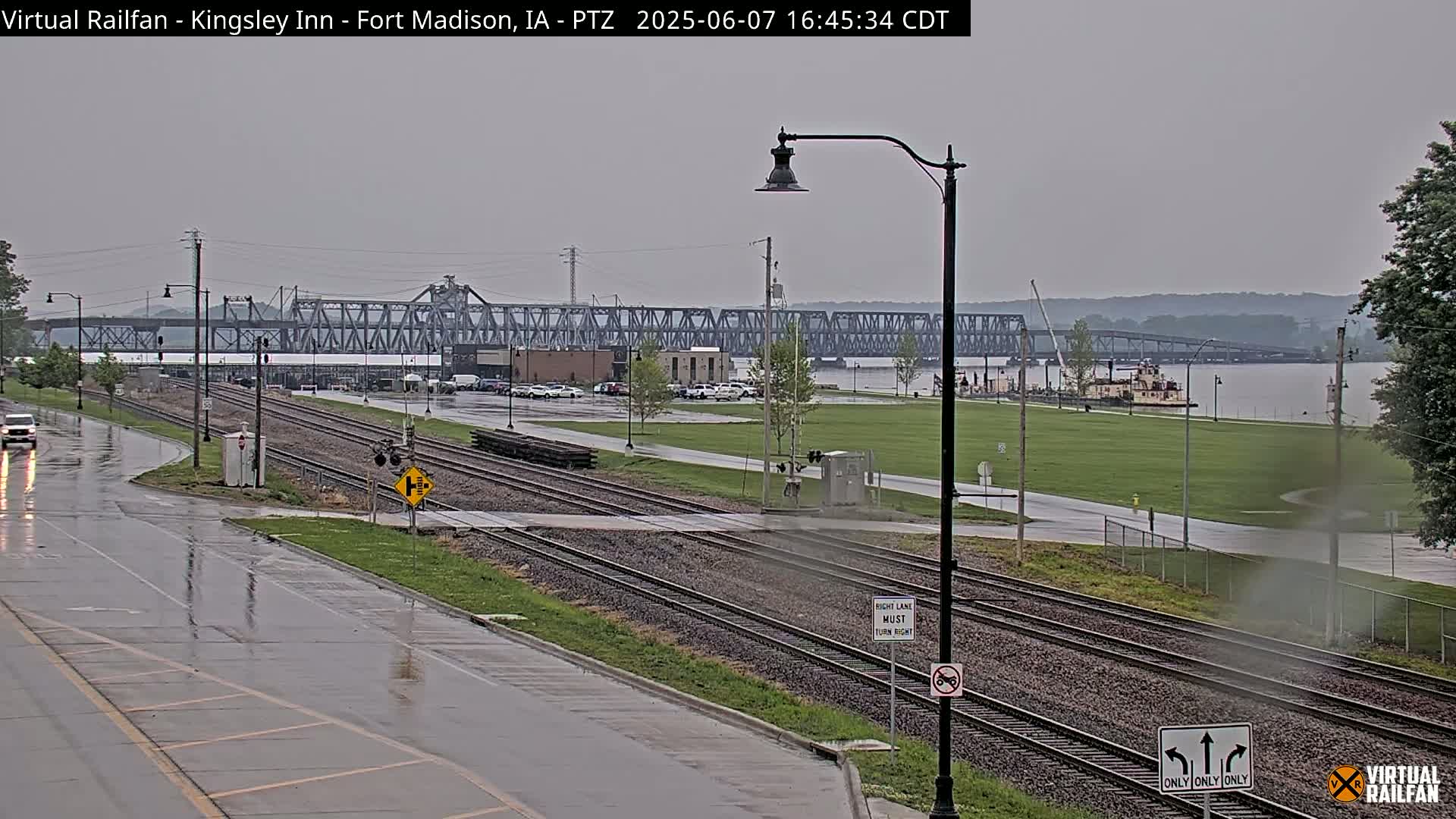 A wet, overcast scene shows railroad tracks intersecting a road next to a river, with a large bridge and a riverboat in the distance.