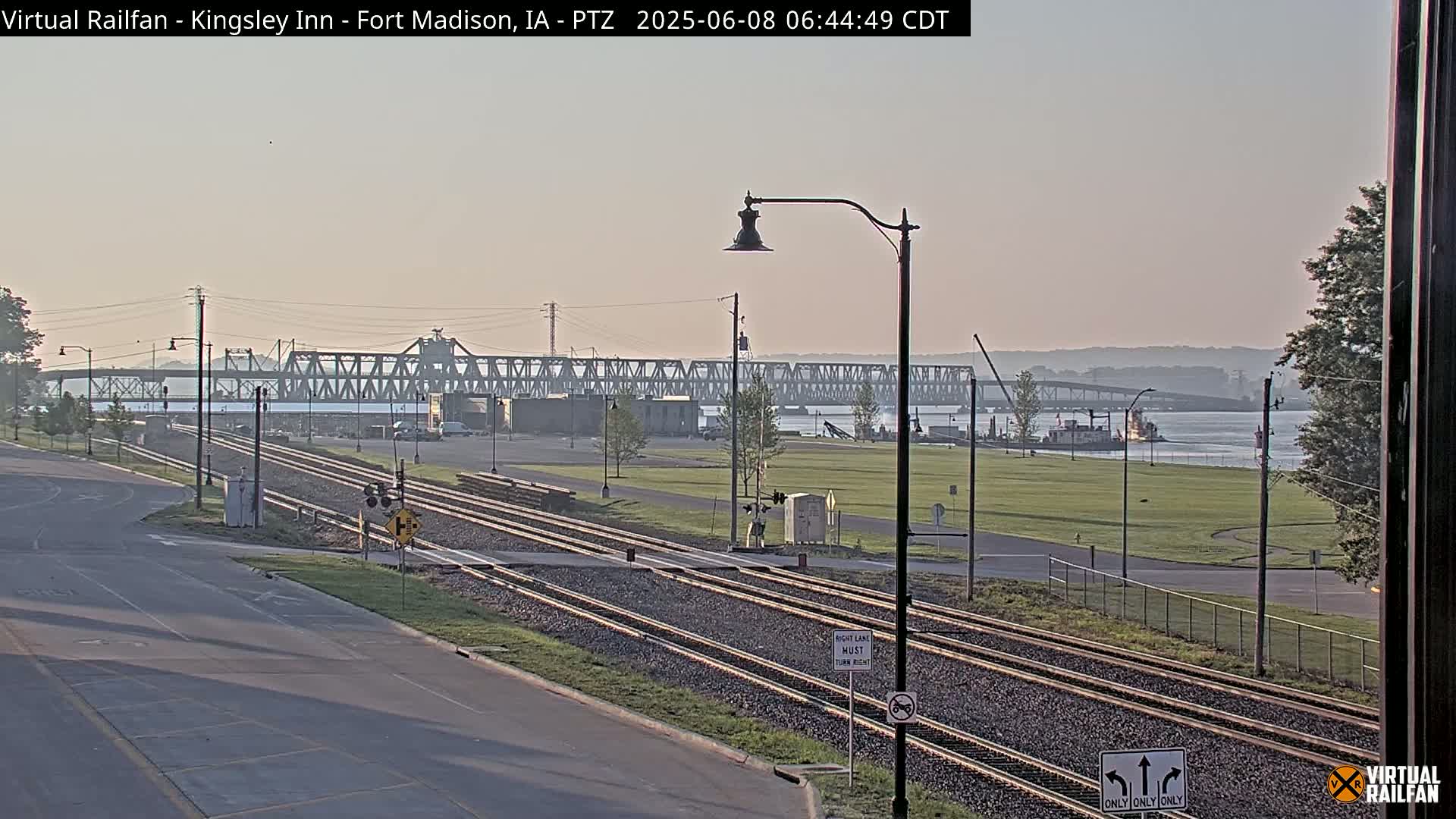 A long, steel bridge spans a river under a hazy, light-colored sky, with railroad tracks and a road in the foreground.