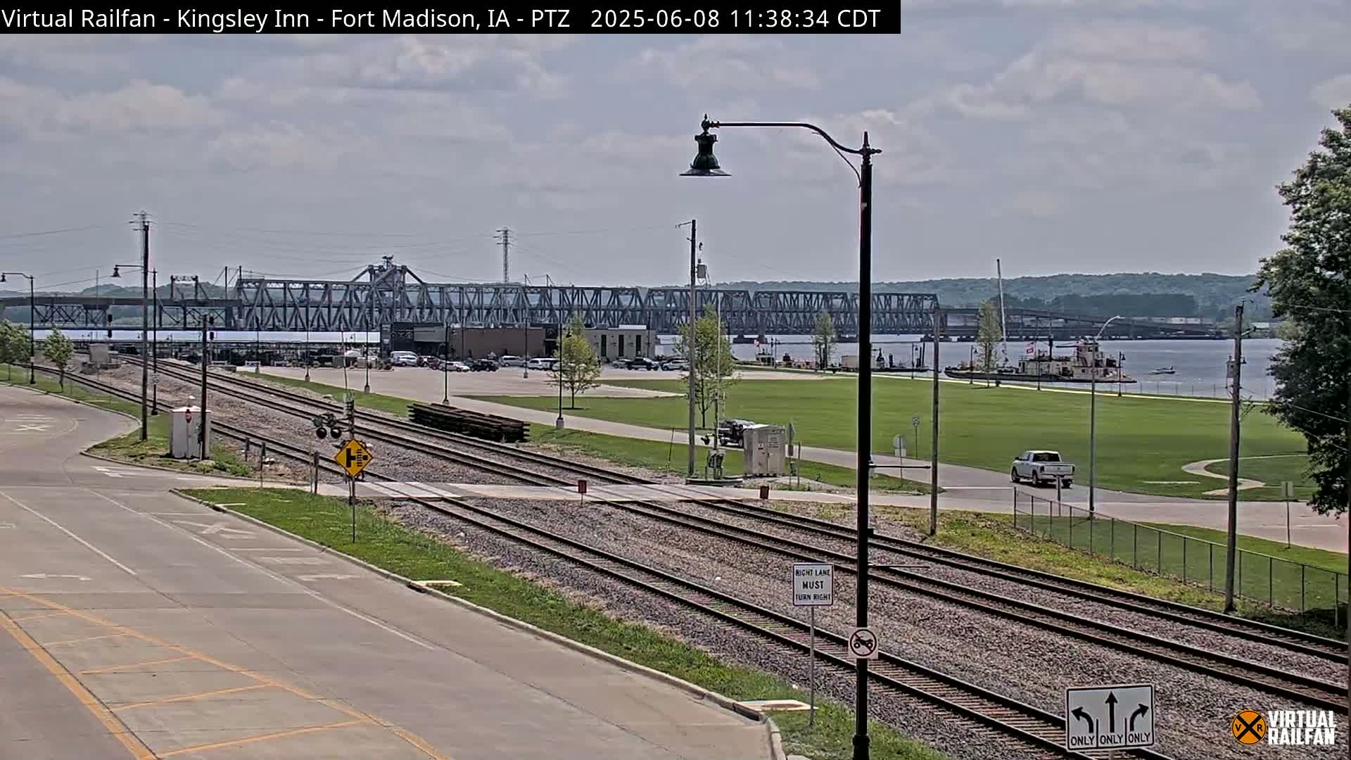 A mostly sunny day shows railroad tracks crossing a road in front of a large metal bridge spanning a river, with a barge visible on the water and a vehicle driving on the road.