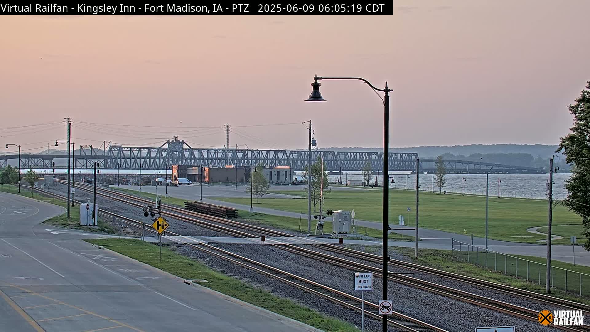 A metal truss bridge spans a river under a pale pink dawn sky, with railroad tracks and a road visible in the foreground.