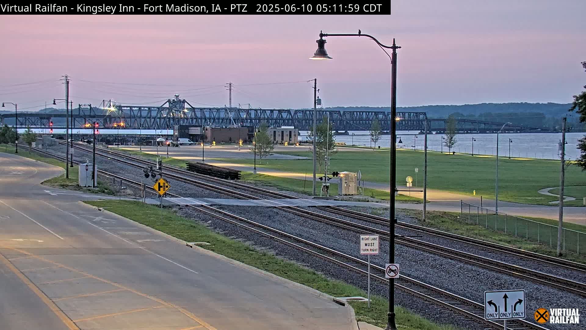 A twilight view of railroad tracks intersecting a road, with a large metal bridge in the background across a body of water.