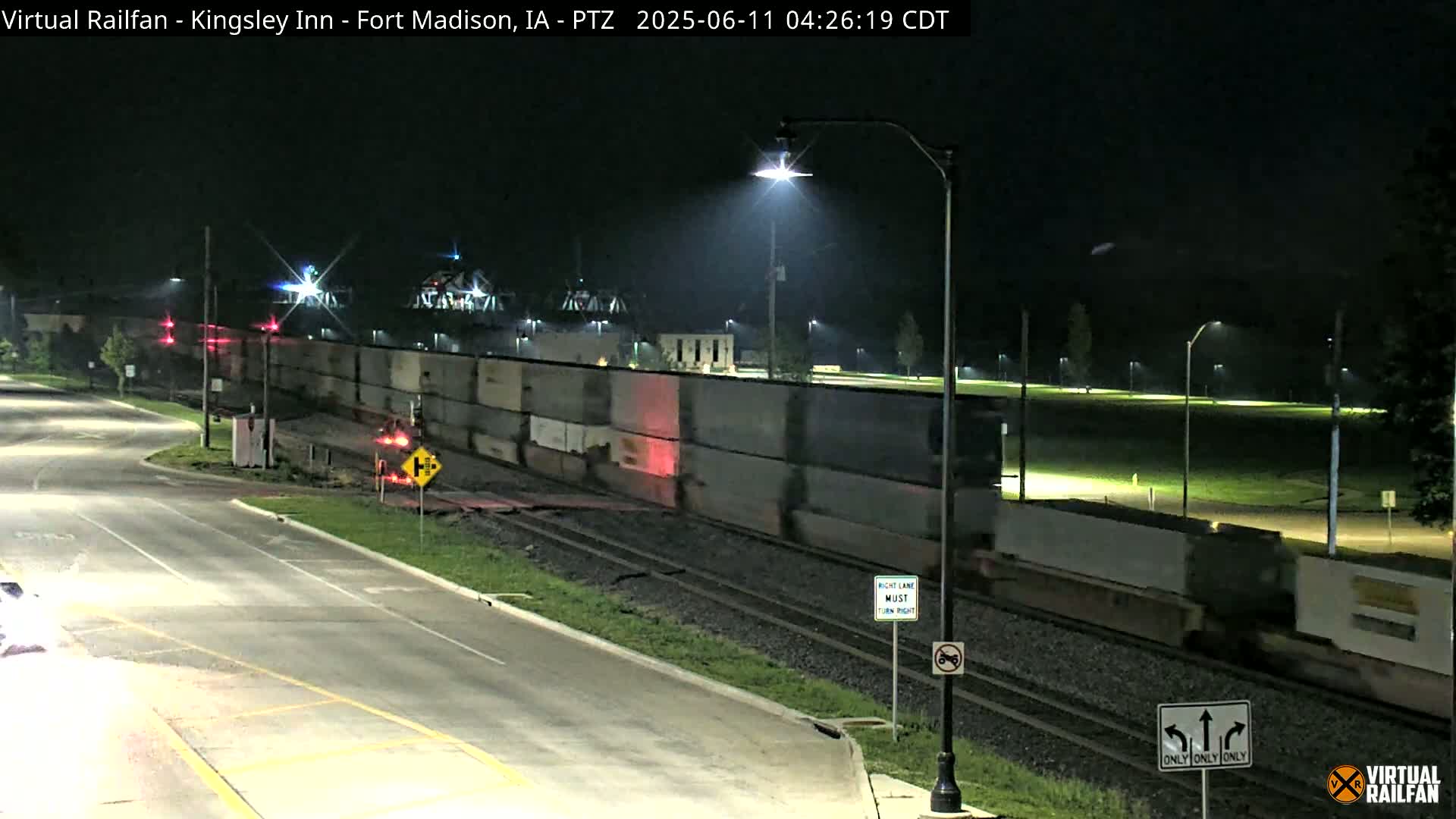 A long freight train passes under a streetlight at night, next to a road with minimal traffic.