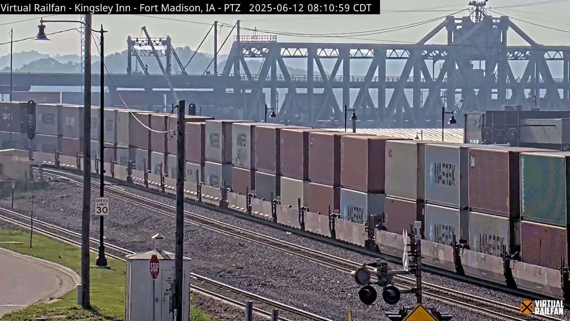 A long freight train carrying numerous shipping containers moves along railroad tracks under a hazy sky, with a large bridge visible in the background.