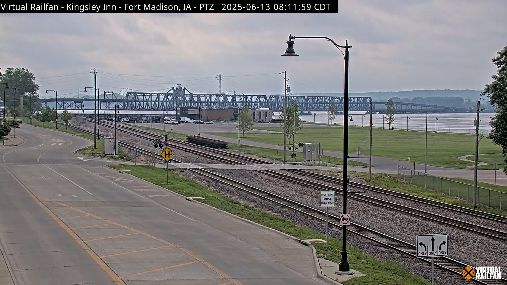 A view of a road and railroad tracks intersecting near a river, with a large metal bridge in the background under an overcast sky.