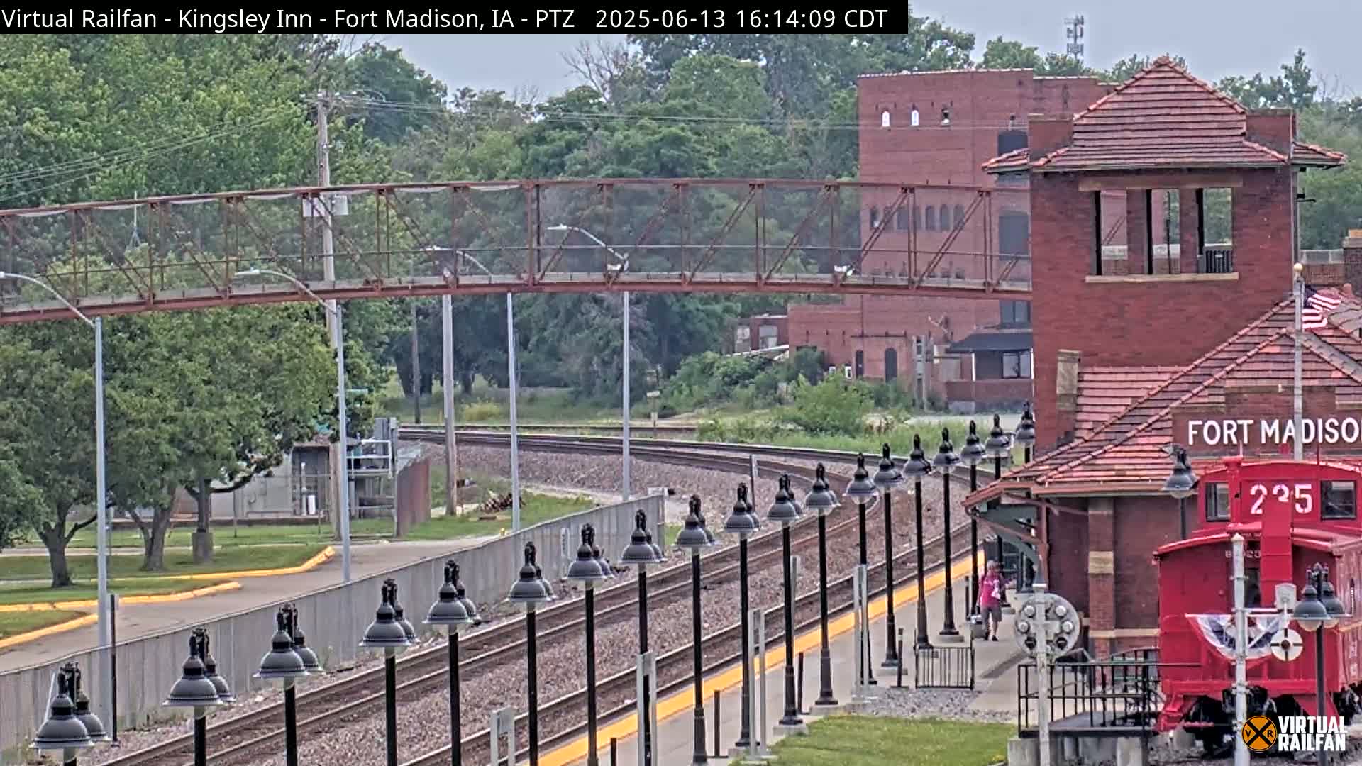 A railway station with a red caboose, a pedestrian bridge, and numerous lamps lines a set of train tracks on an overcast day.