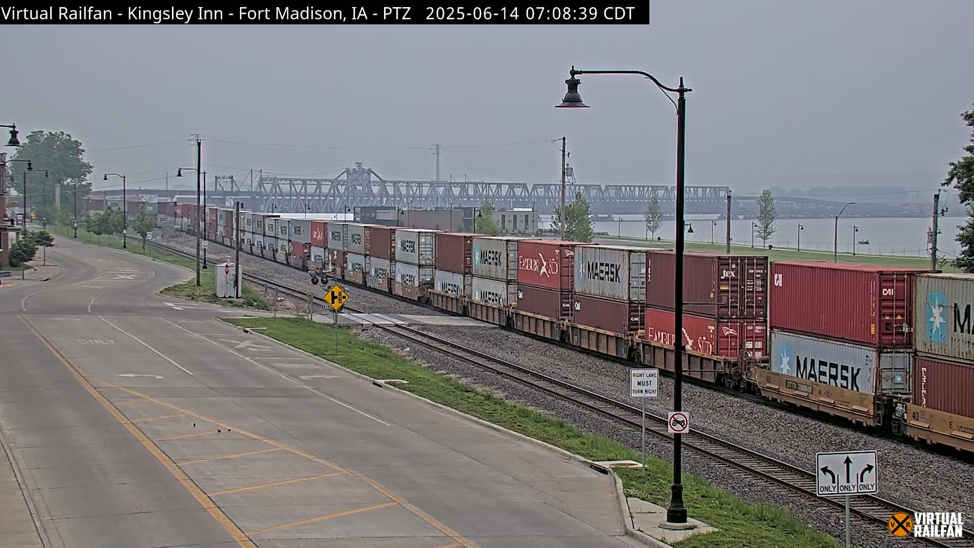 A long freight train carrying numerous cargo containers travels along railroad tracks next to a road on a hazy day, with a river and bridge visible in the background.