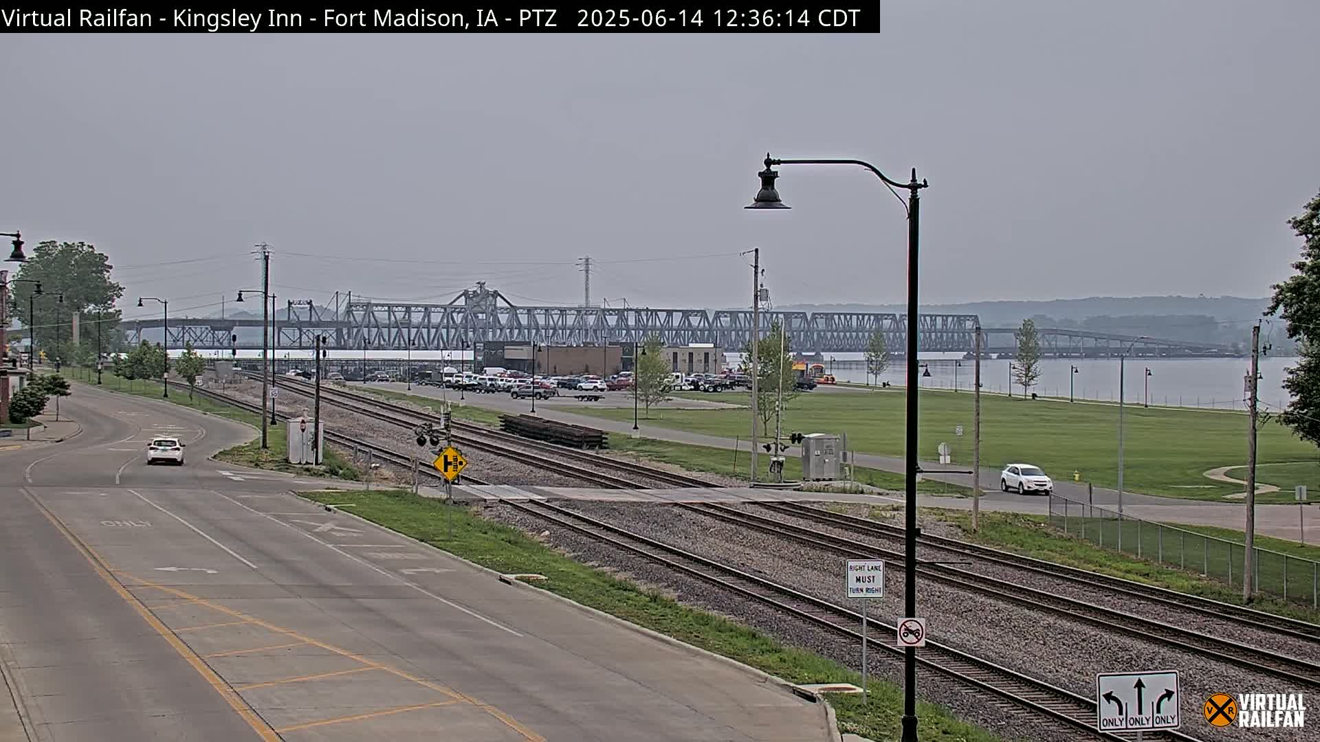 A long road with railroad tracks runs alongside a river, under an overcast sky, with a large metal bridge in the distance and a few vehicles on the road.