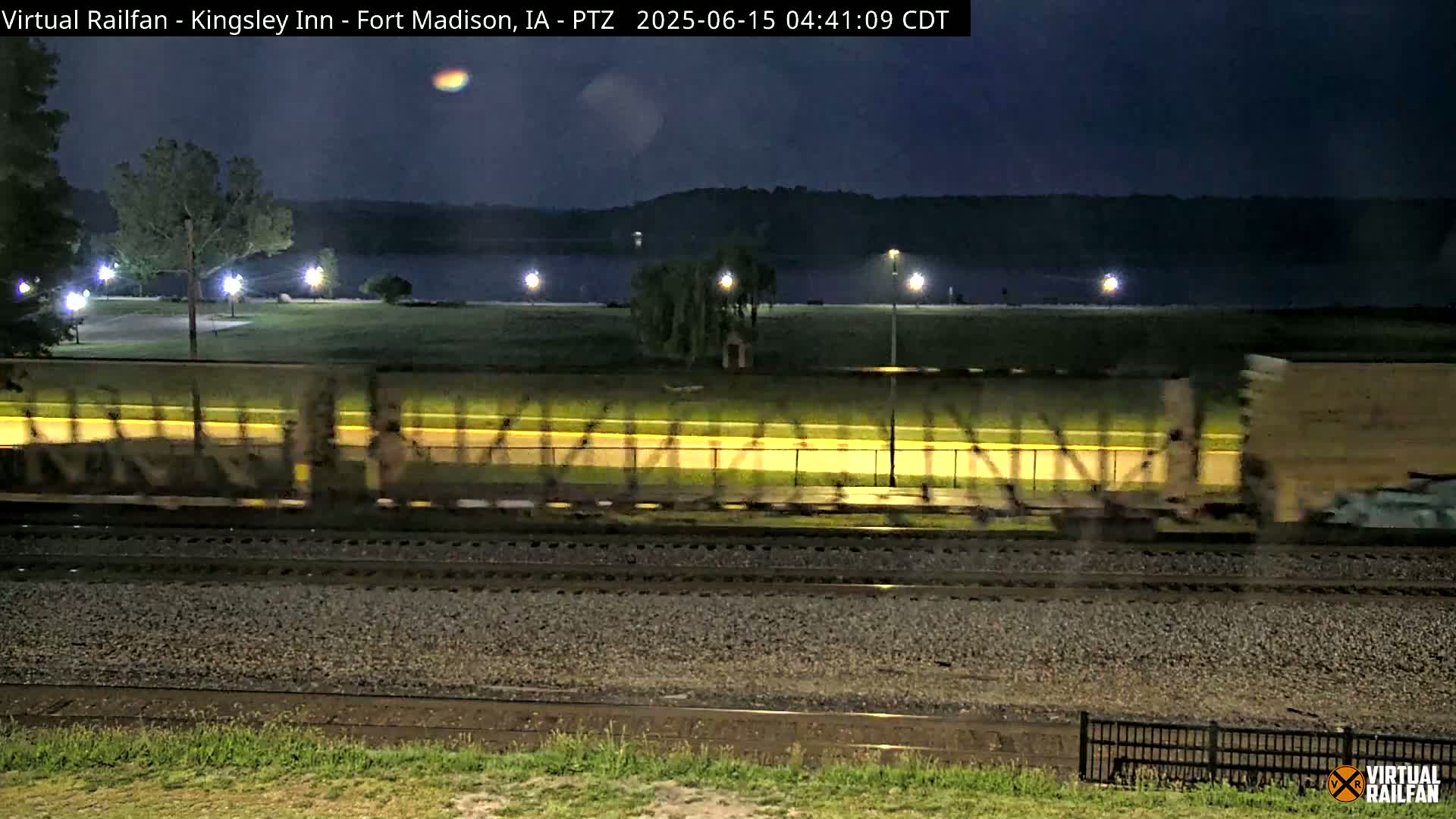 A long freight train moves along railroad tracks at night next to a park lit by streetlights beside a dark body of water.