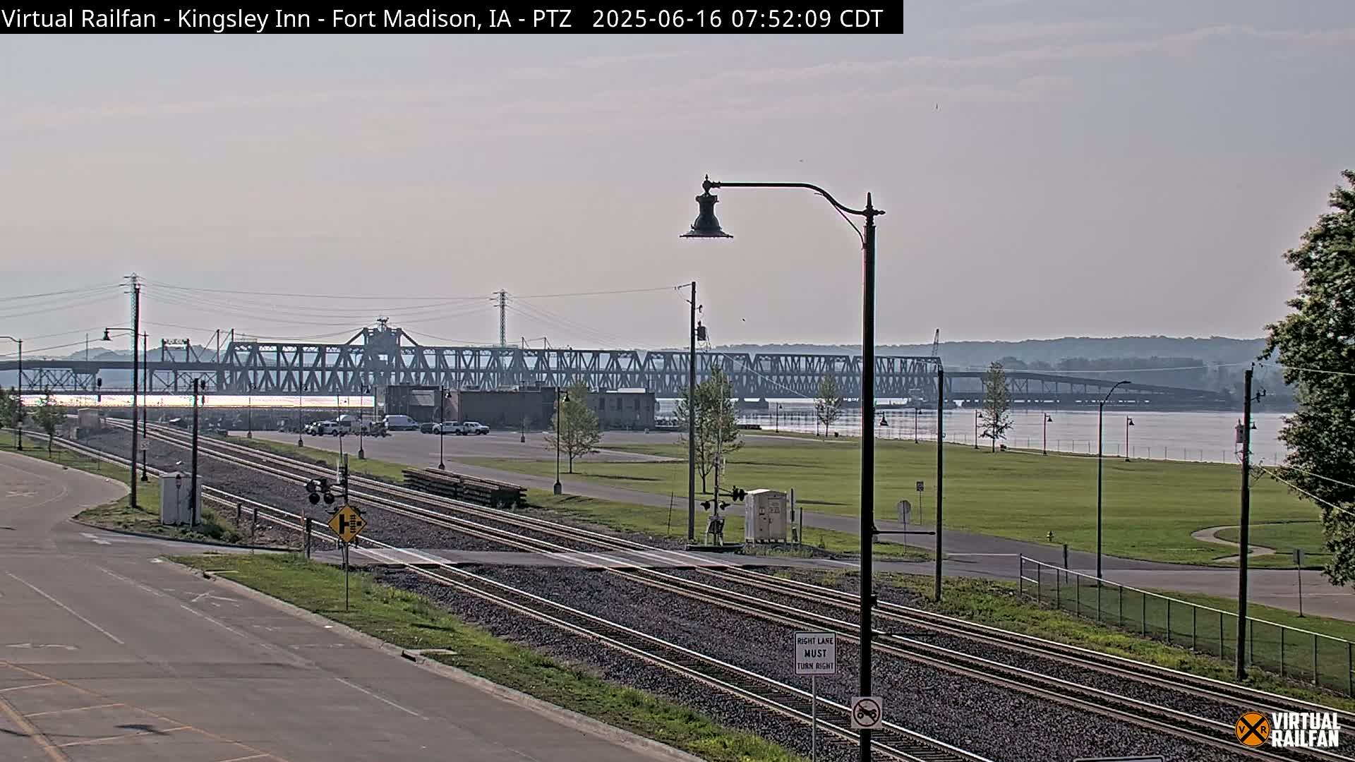 A view of railroad tracks leading to a metal bridge over a wide river under a partly cloudy sky.