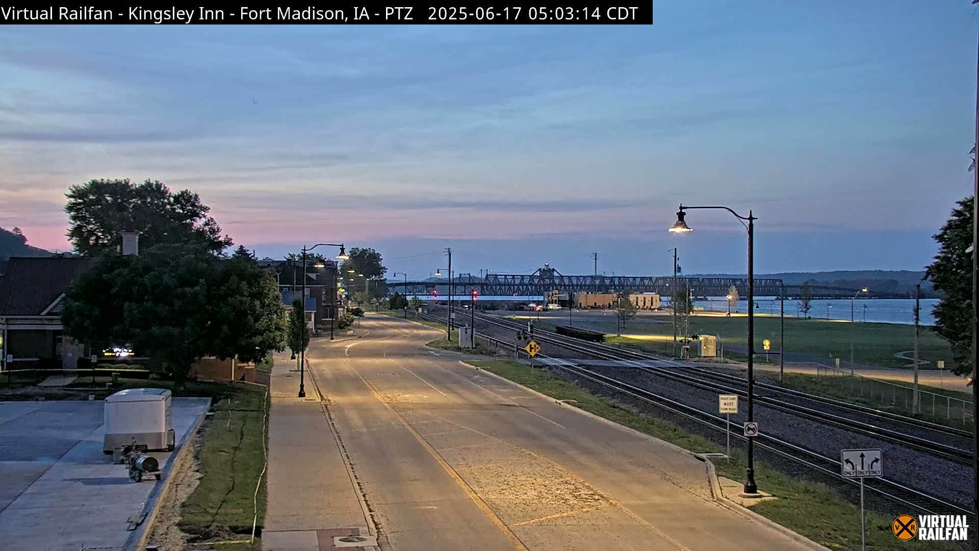 A mostly empty, two-lane road at twilight, lined with streetlights and bordered by railroad tracks, leads toward a river and a bridge under a light-pink and blue sky.