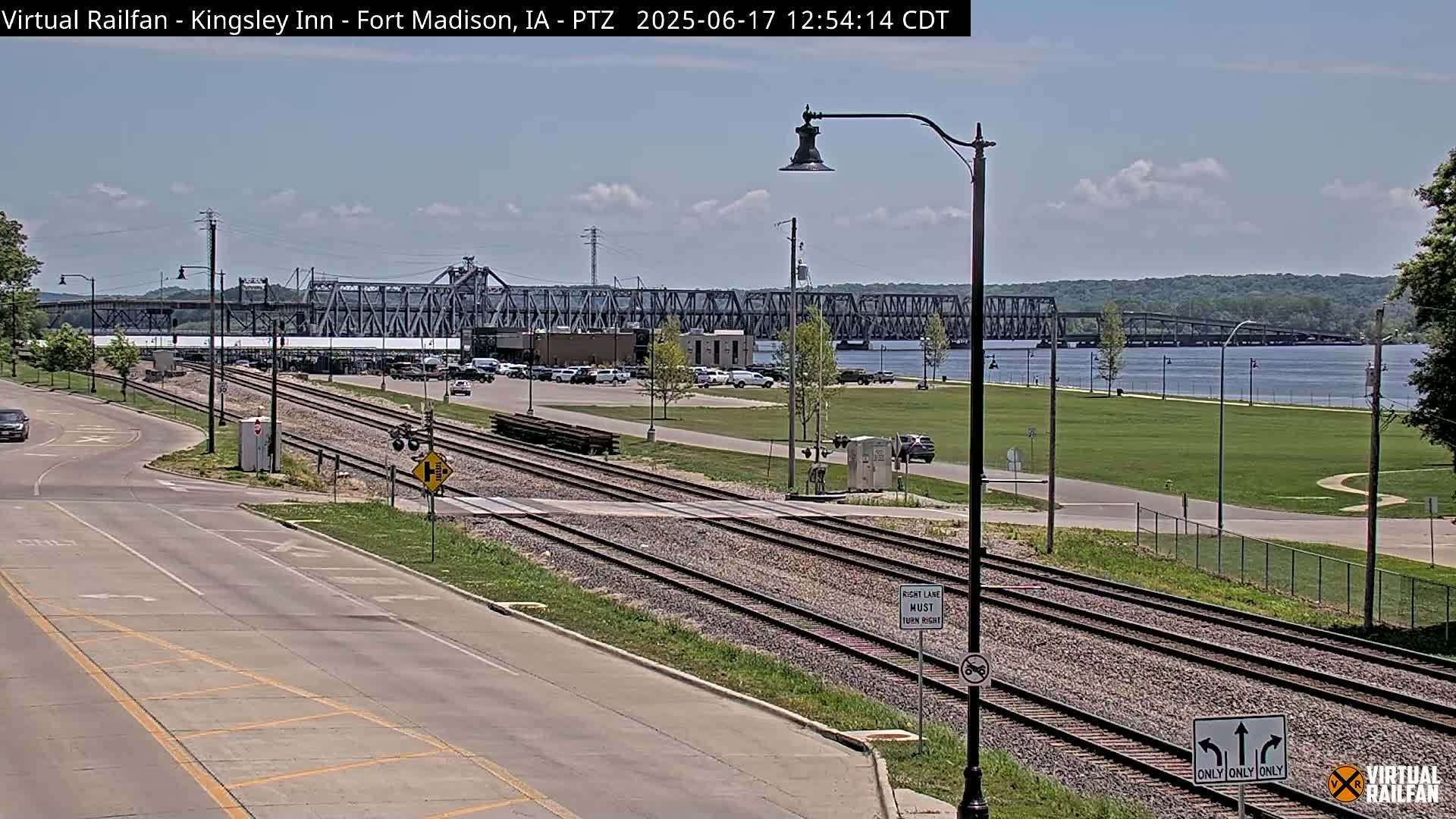A long view of a river, a bridge, train tracks, a road, and a grassy area under a partly cloudy sky.