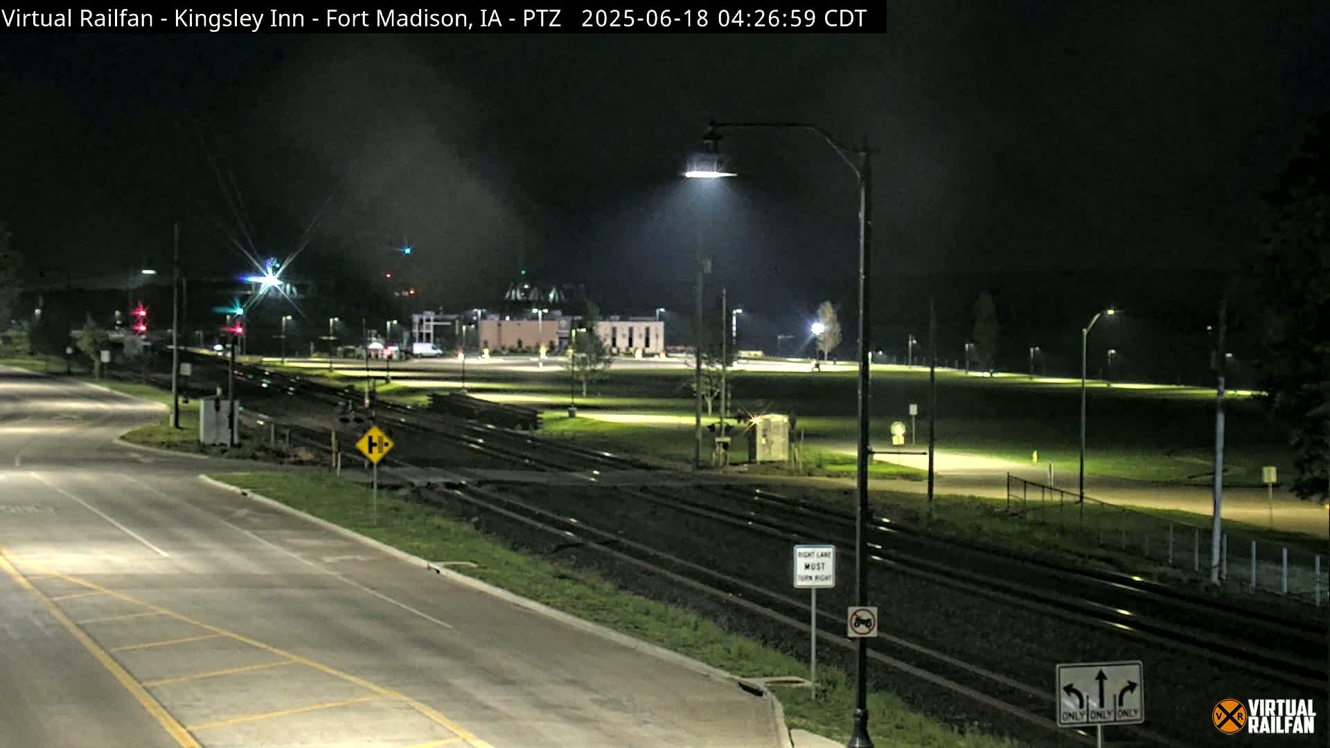 A nighttime view of a roadway intersecting with railroad tracks, passing near a building and a grassy area illuminated by streetlights.