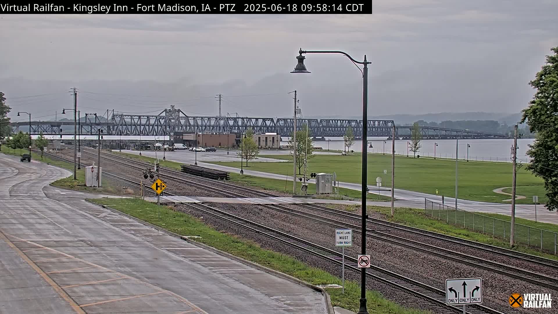 A view of railroad tracks and a road leading to a large metal bridge over a river, under overcast skies.
