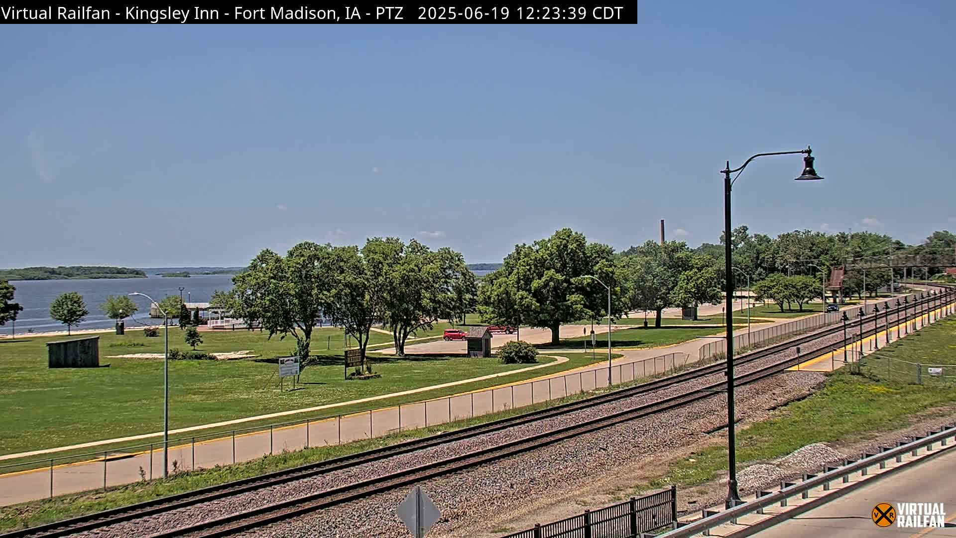 A sunny day shows a park with green grass, trees, a roadway, and railroad tracks beside a body of water.
