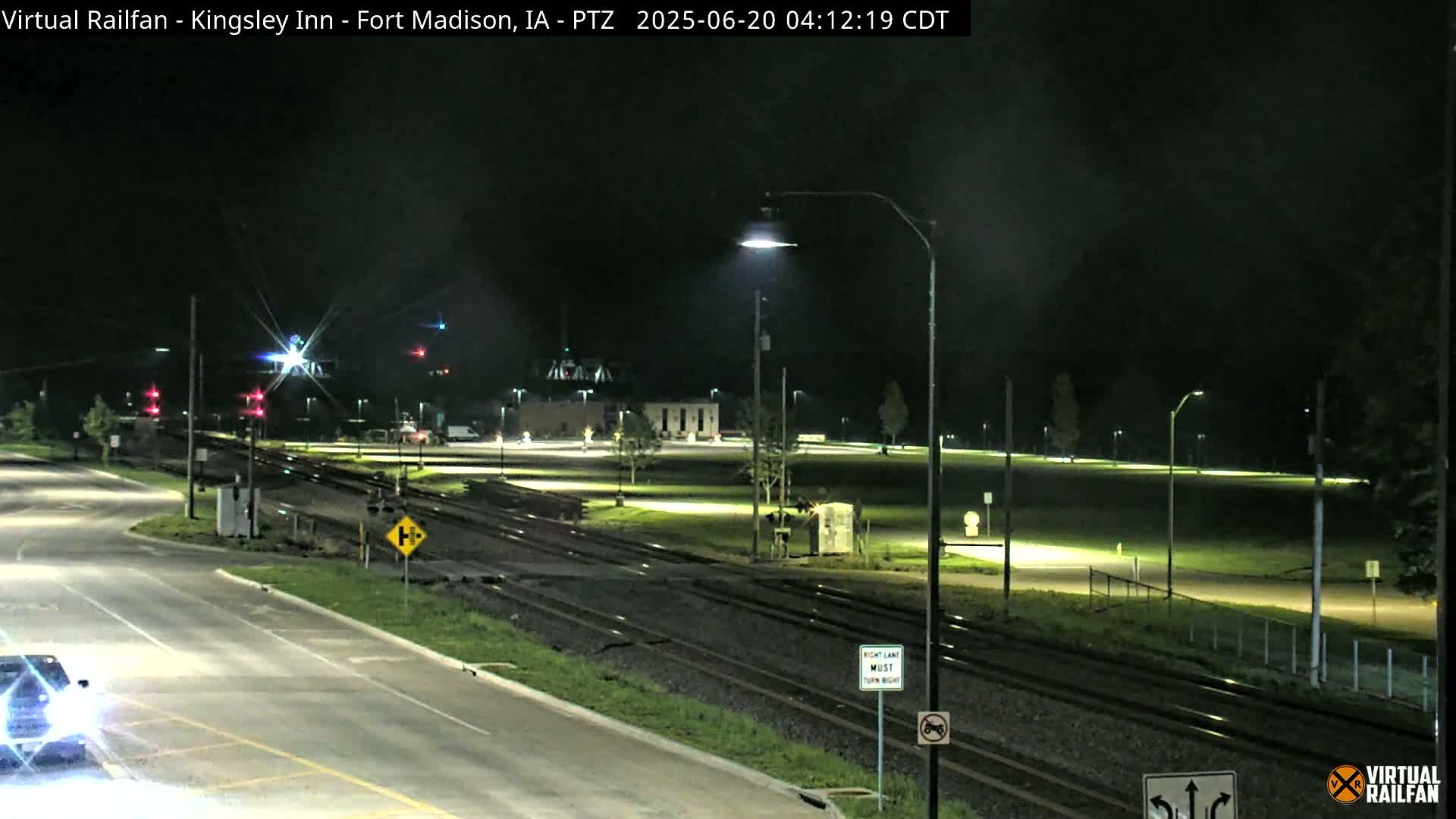 A nighttime view of a road intersection near railroad tracks, with a few cars visible and streetlights illuminating a grassy area.