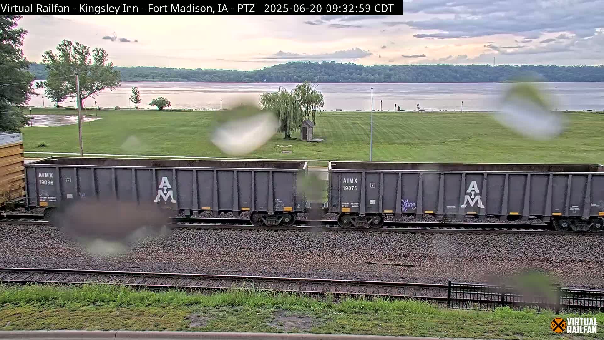 Two gray rail cars sit on train tracks overlooking a calm river and grassy field under an overcast sky.