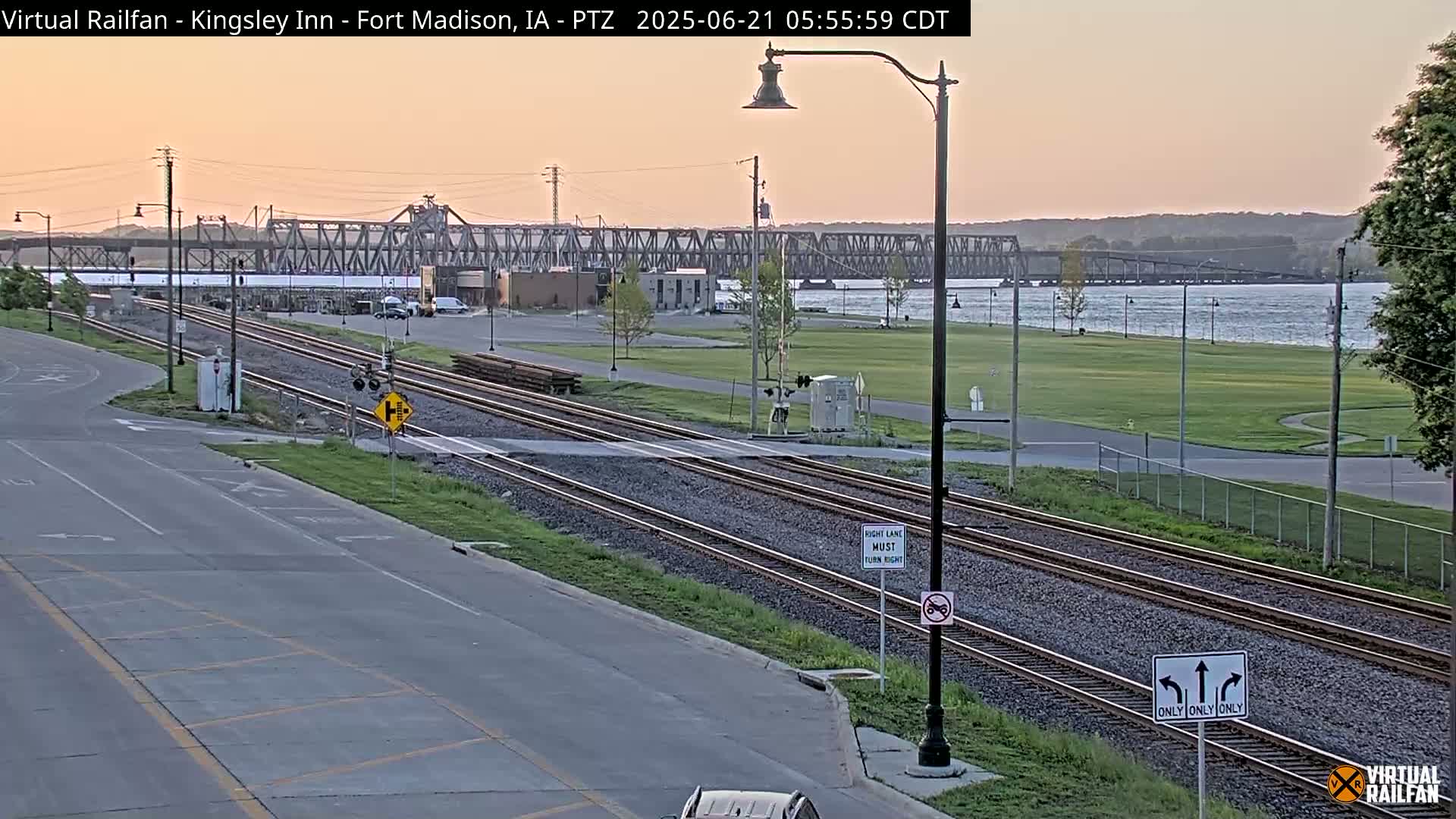 A twilight view of train tracks intersecting a road, with a river and bridge in the background under a clear, pinkish sky.