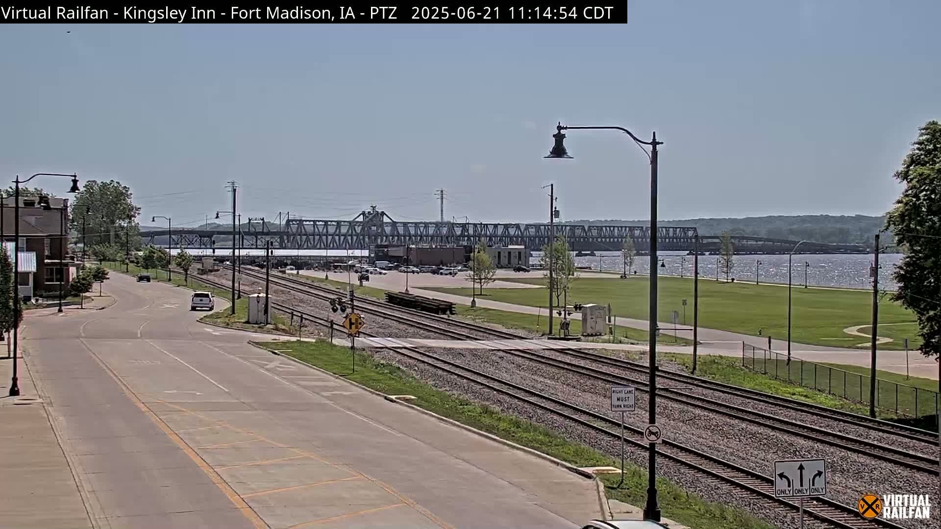 A sunny day shows a view of a street with railroad tracks crossing it, leading to a river with a large bridge in the distance.