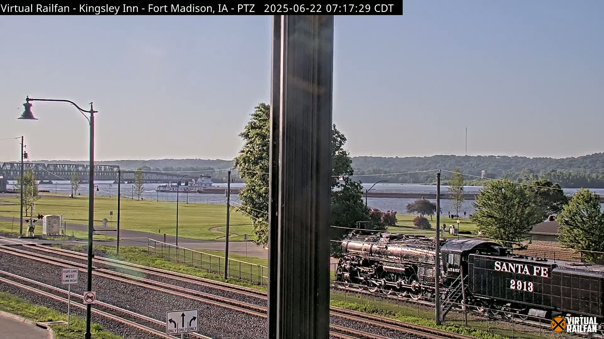 A black steam locomotive is parked on train tracks near a river under a mostly clear sky, with a bridge and barge visible in the background.
