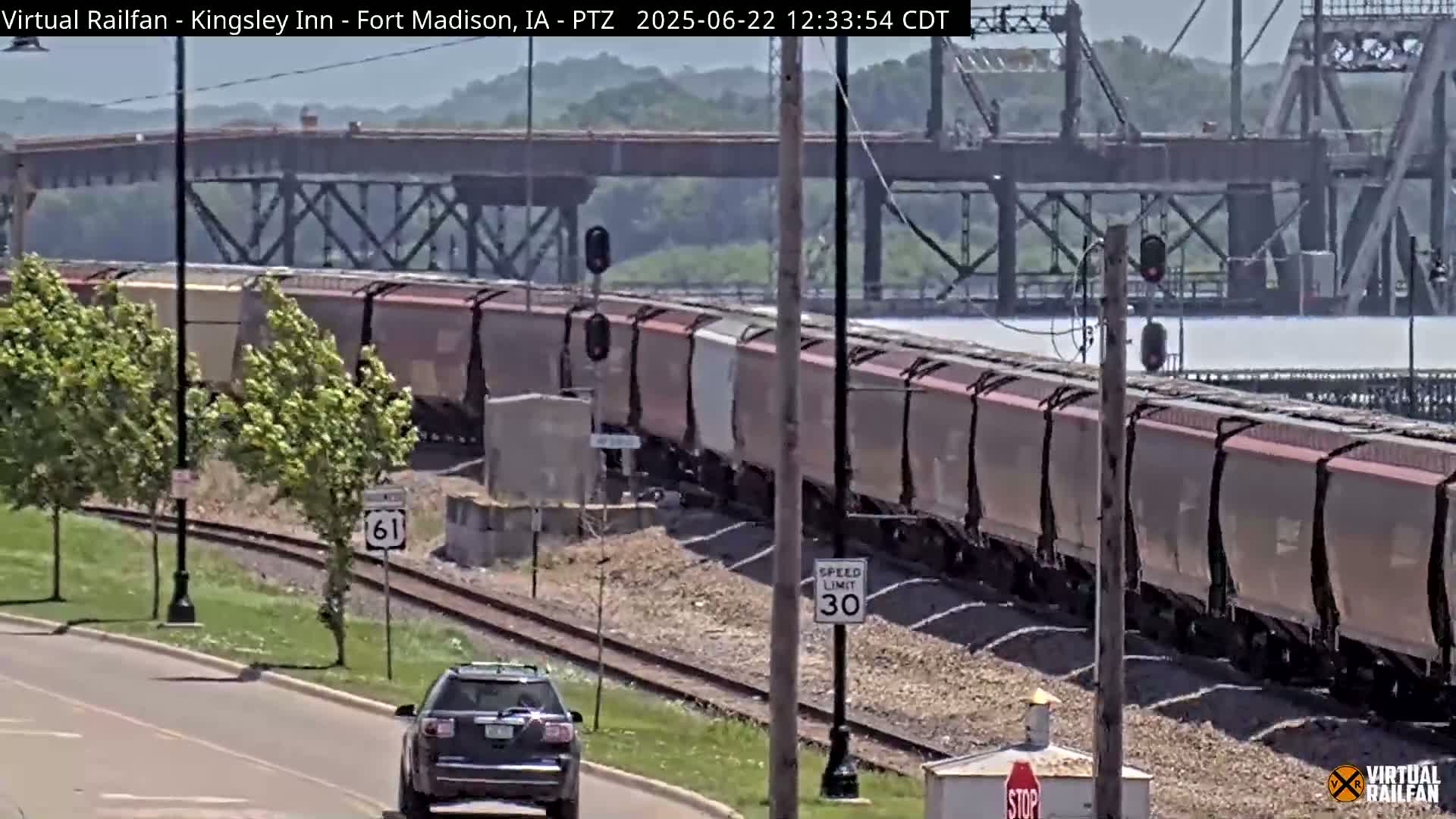 A long freight train passes under a bridge on a sunny day near a road with a car and trees.