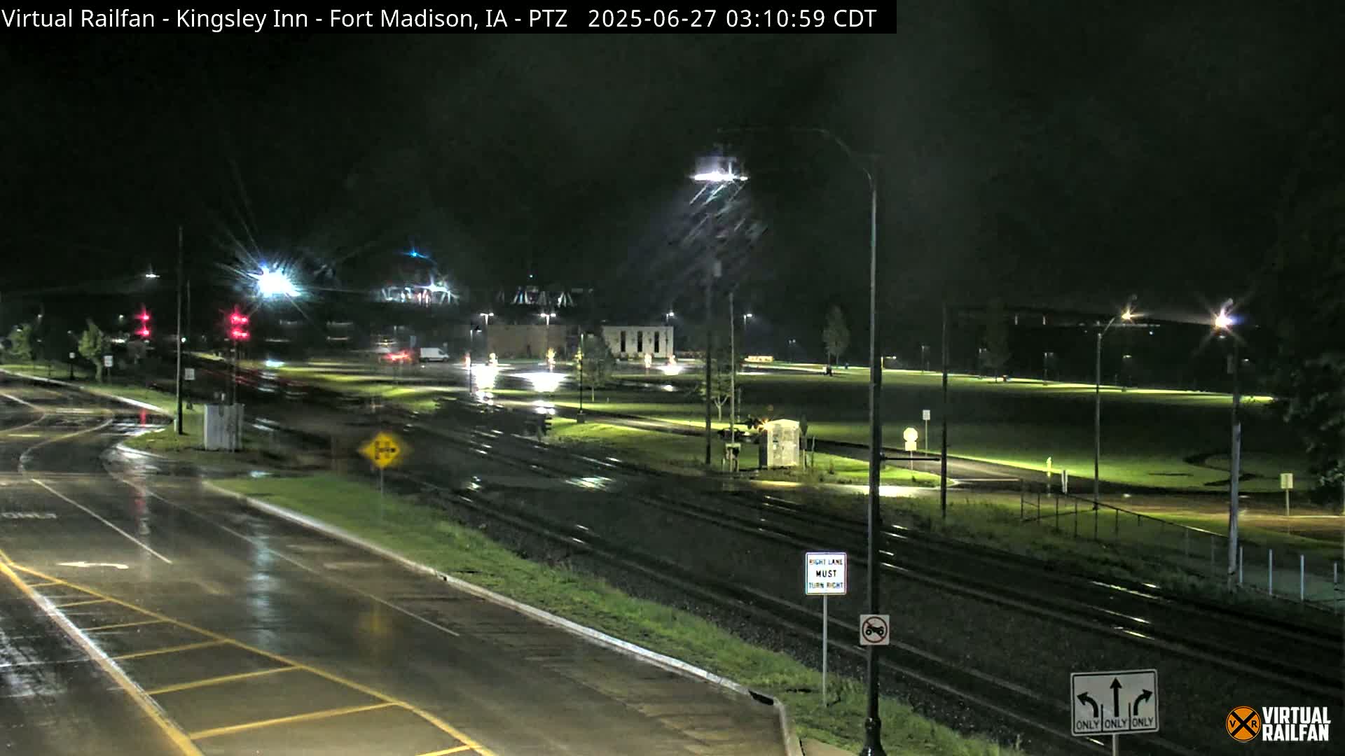 A rain-slicked nighttime intersection shows railroad tracks intersecting roads, with streetlights illuminating wet pavement and sparse traffic.