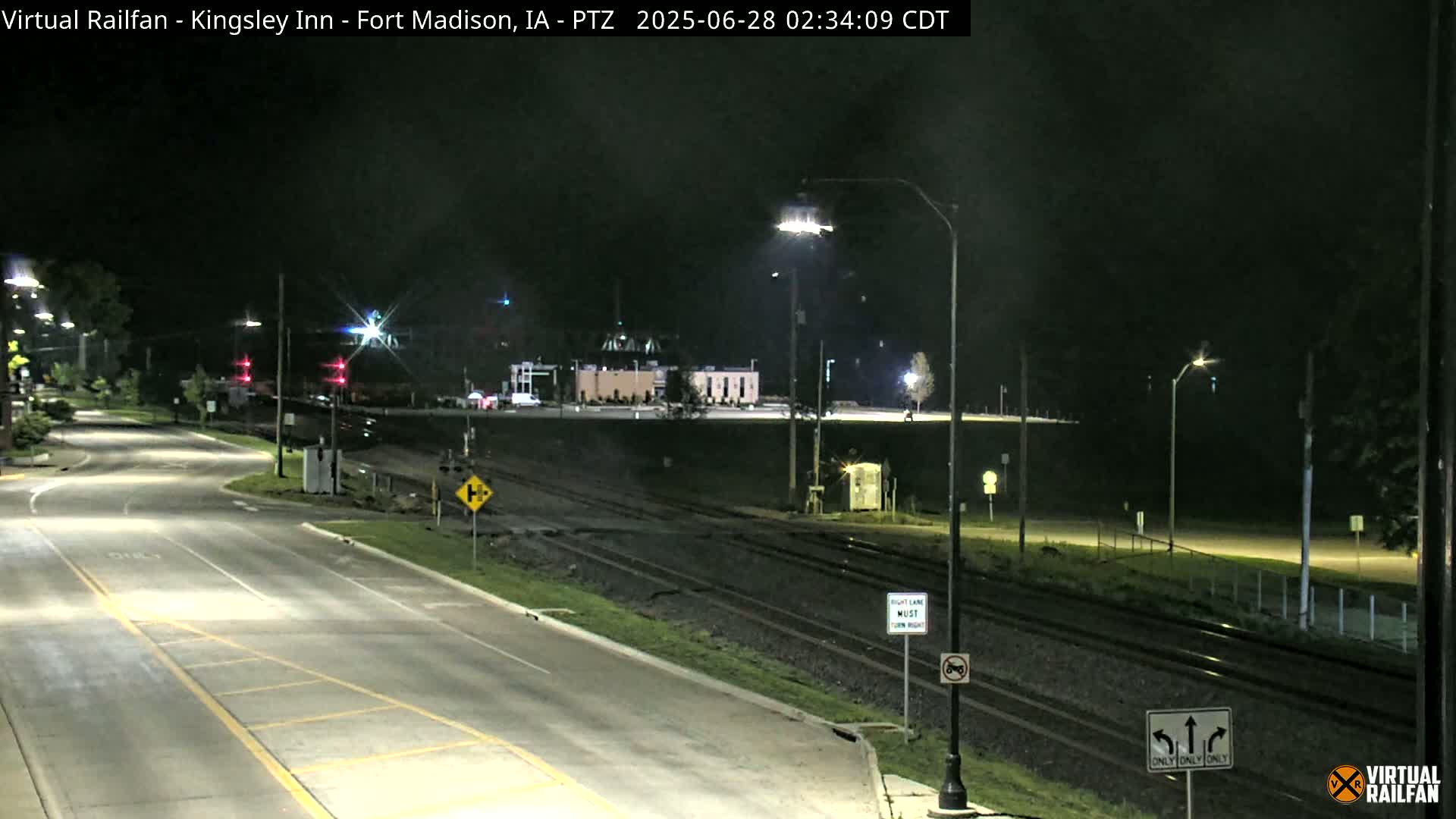 A nighttime view of a road intersecting with train tracks, showing a building in the distance under clear skies.