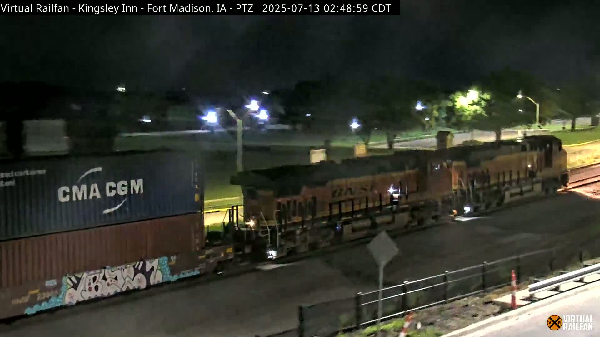 At night, a freight train with a graffiti-covered container car passes by under illuminated streetlights.