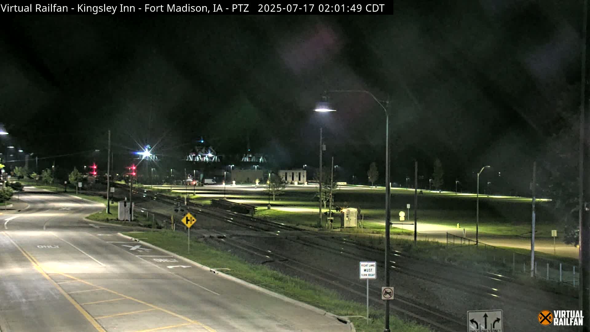 A nighttime view of a roadway intersecting with railroad tracks, featuring well-lit streets and surrounding grassy areas under a dark, clear sky.