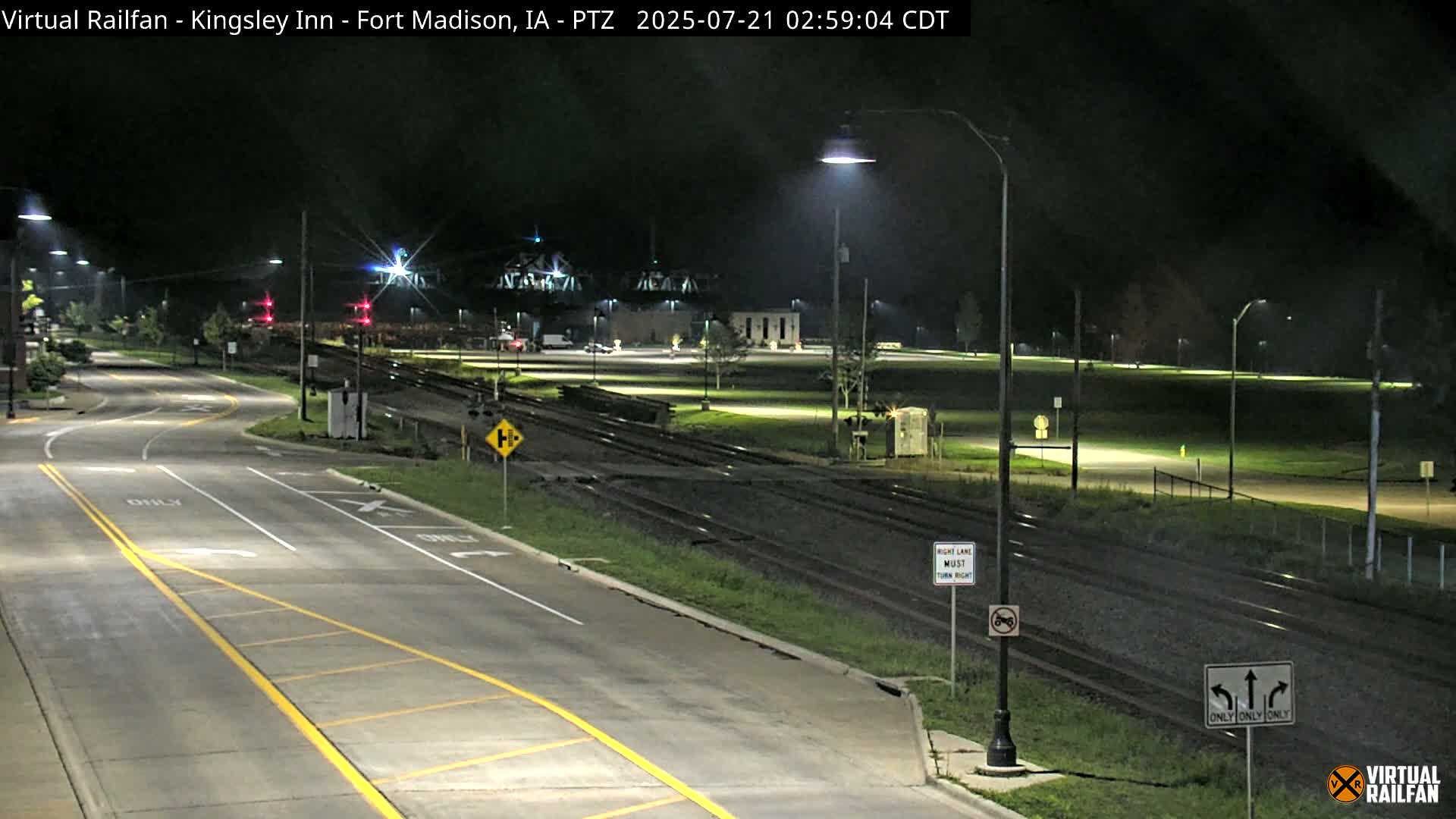 A nighttime view of a paved road intersection with railroad tracks nearby, illuminated by streetlights.