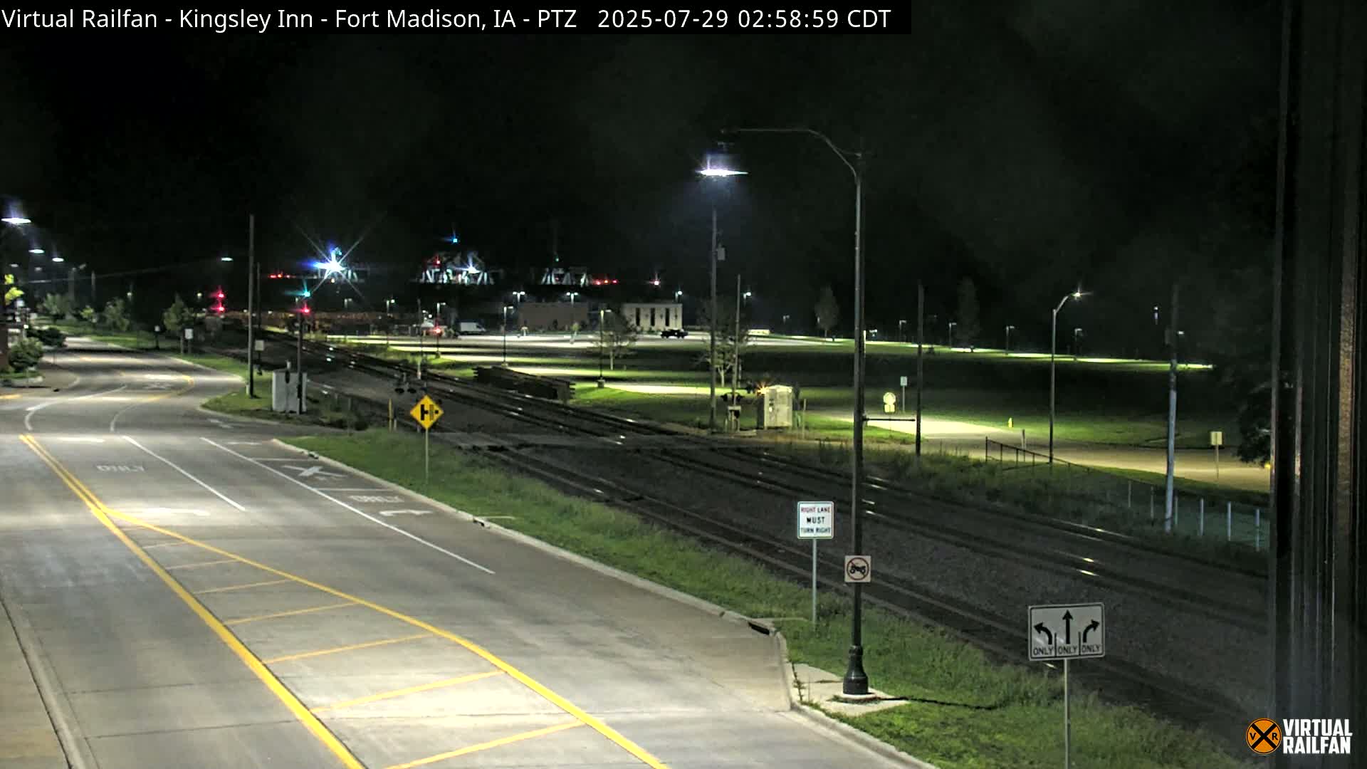 A nighttime view of a paved road intersecting with railroad tracks, under well-lit streetlights, with a grassy area and buildings in the background.