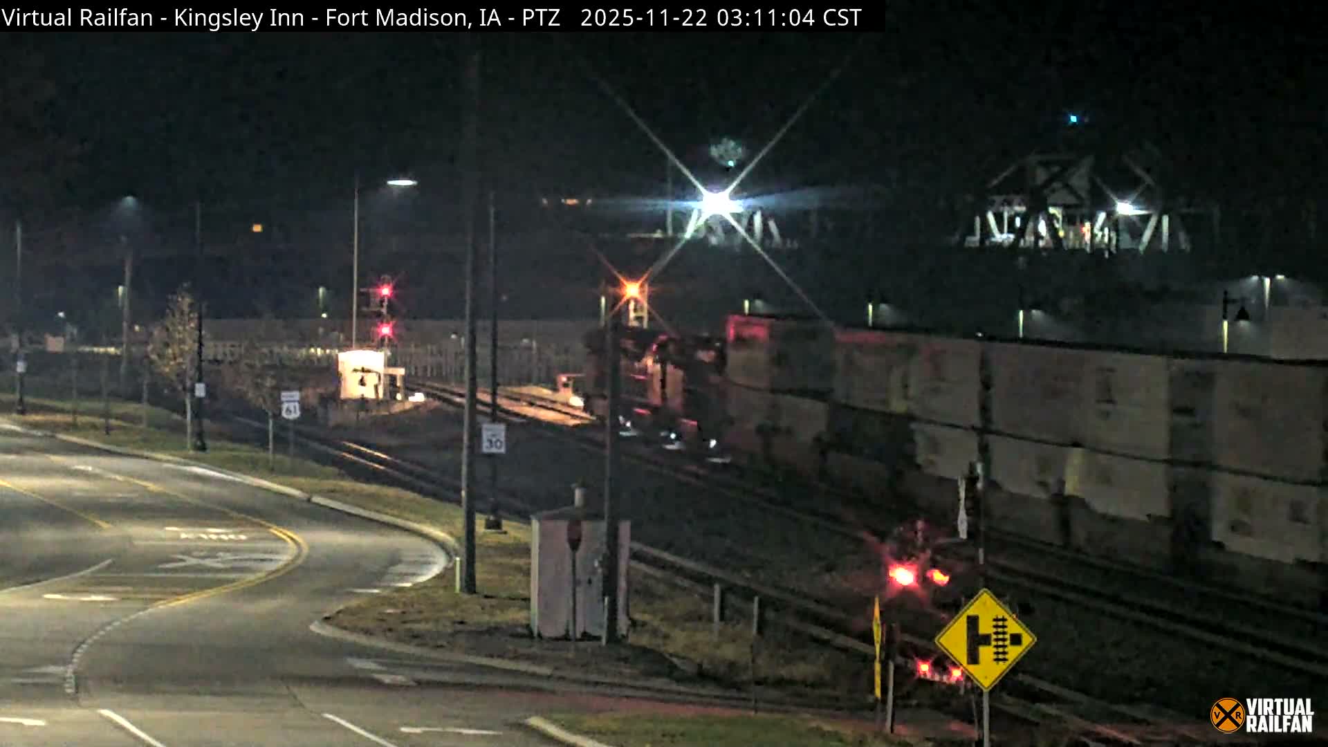 A train with illuminated headlights and freight cars is visible on multiple tracks, passing alongside a curved road with streetlights and red signals, during a clear night.