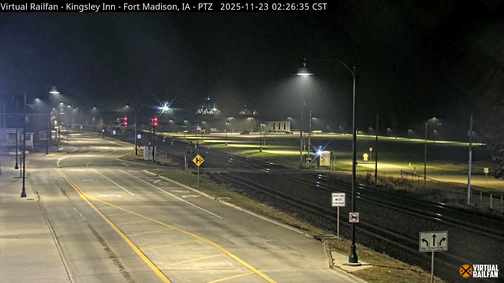 A clear night view displays a multi-lane road and multiple railroad tracks illuminated by numerous streetlights, with buildings and a large bridge visible in the distance.